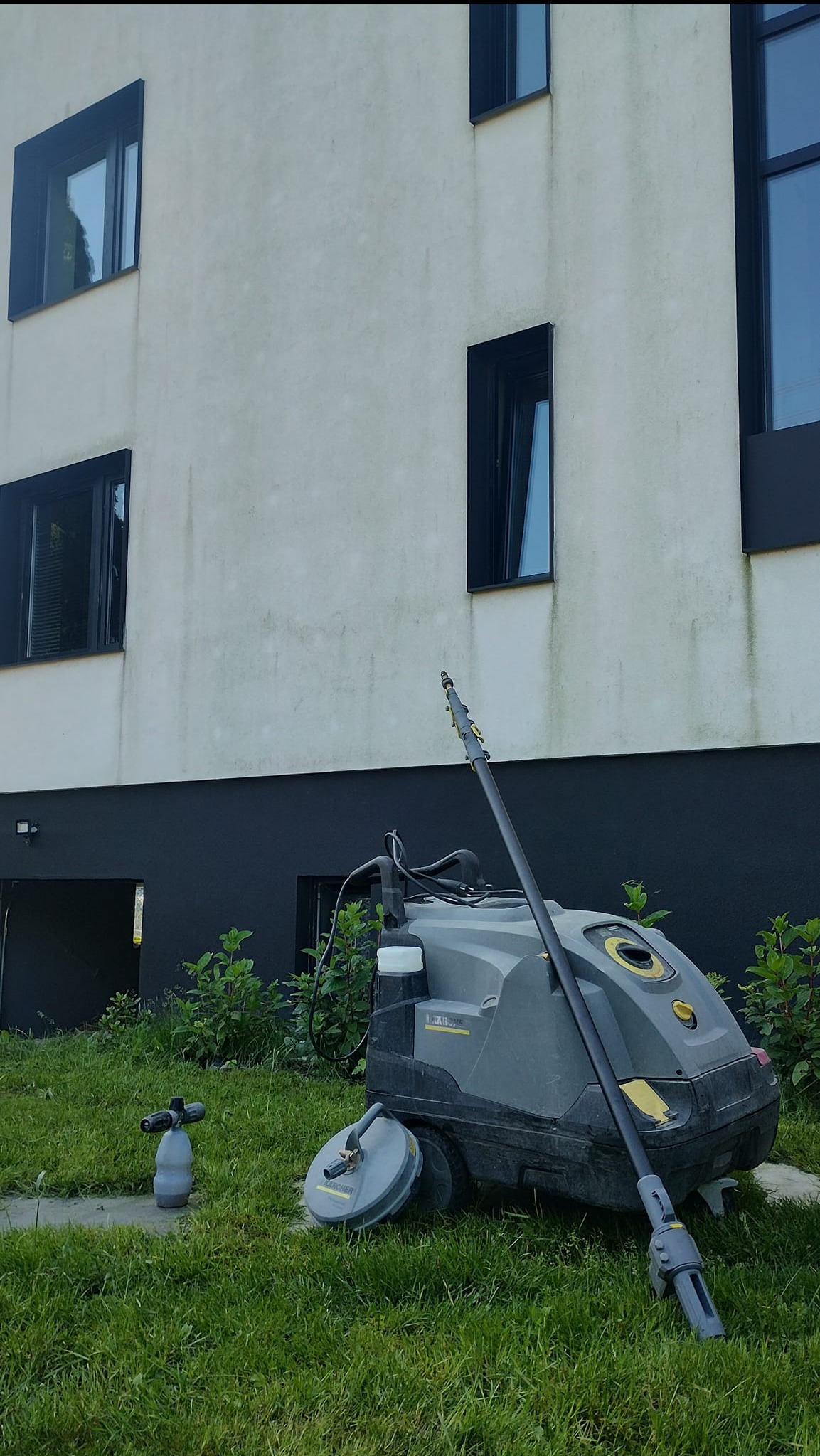 Urządzenie wysokociśnieniowe Karcher na trawie przed budynkiem z zabrudzoną elewacją. Widoczna dysza i przystawka do mycia powierzchni płaskich. Przygotowanie do czyszczenia.