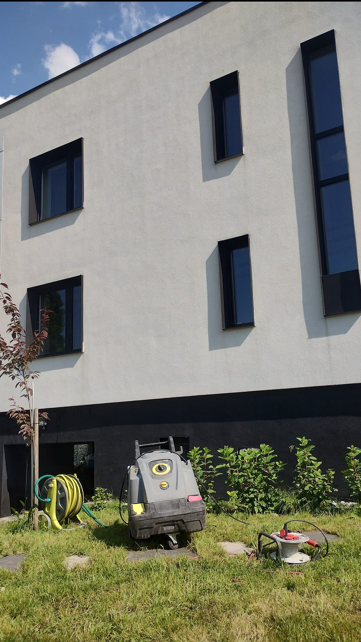Szary, nowoczesny budynek z ciemnymi oknami. Na trawie stoi myjka ciśnieniowa Kärcher i zwinięty wąż ogrodowy, gotowe do czyszczenia elewacji.