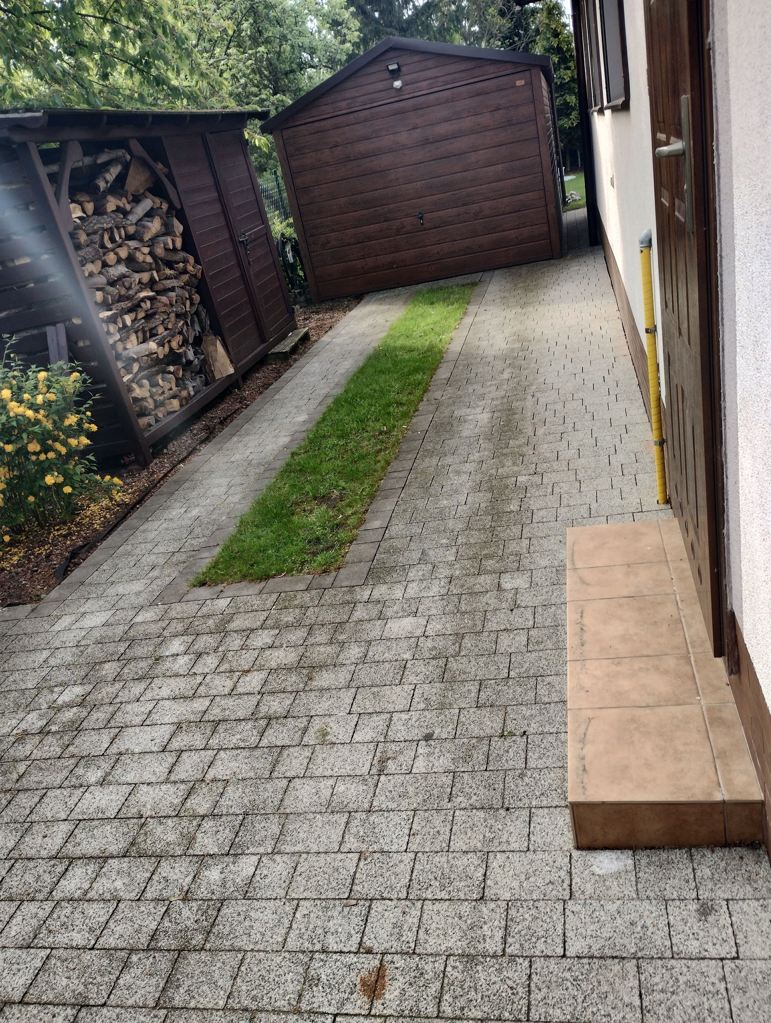 Zewnętrzna kostka brukowa prowadząca do drzwi domu, z widocznymi zabrudzeniami i mchem. Obok drewutnia i garaż. W tle zielona roślinność.