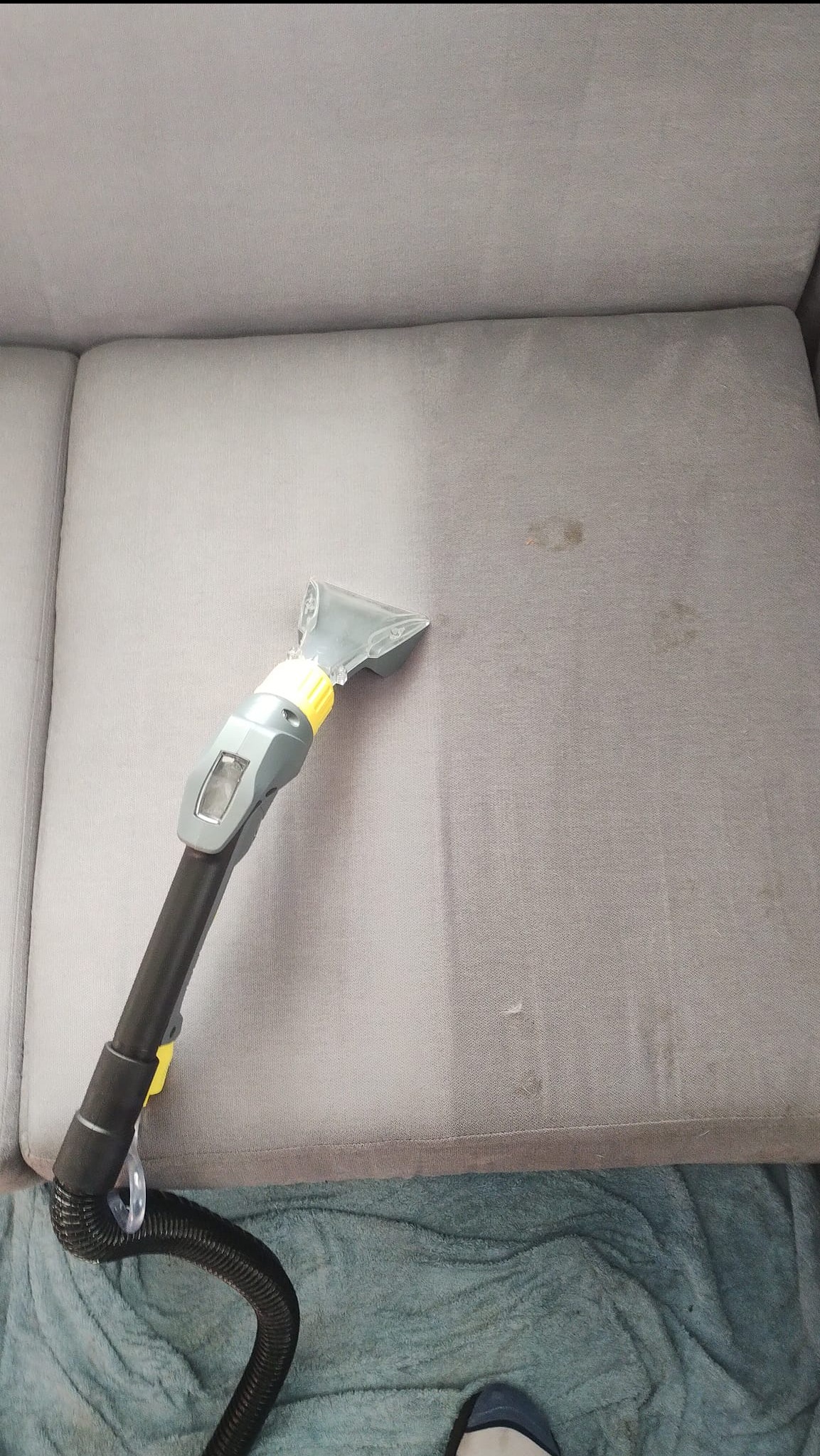 Czyszczenie tapicerki kanapy parownicą, widoczne różnice w czystości fragmentu po czyszczeniu. W tle niebieski koc i fragment buta.