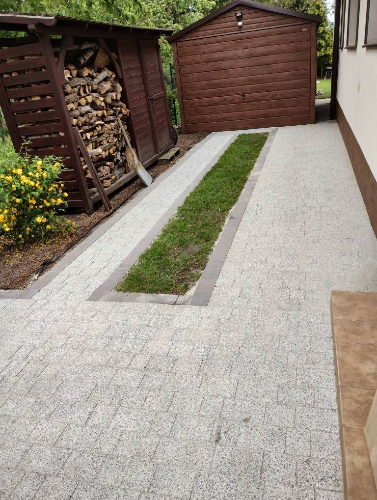 Jasnoszara kostka brukowa z wstawkami trawnika, prowadząca do drewnianego garażu i drewutni. Ułożona kostka z regularnym wzorem, widoczne detale wykończenia.