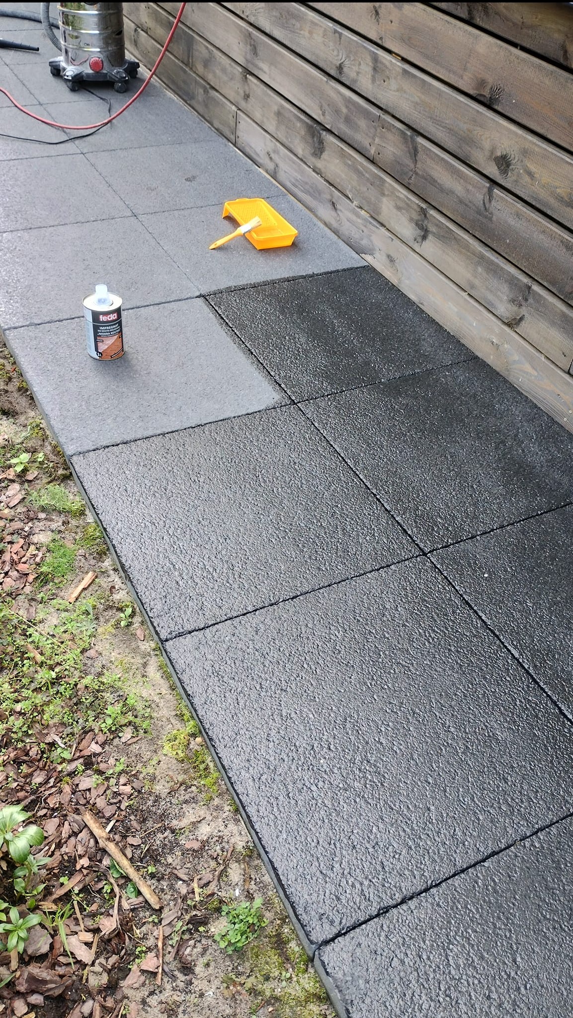 Impregnacja kostki brukowej na zewnątrz. Częściowo zaimpregnowana kostka, pędzel w kuwecie, preparat impregnujący. Widoczne drewniane elementy wykończeniowe.