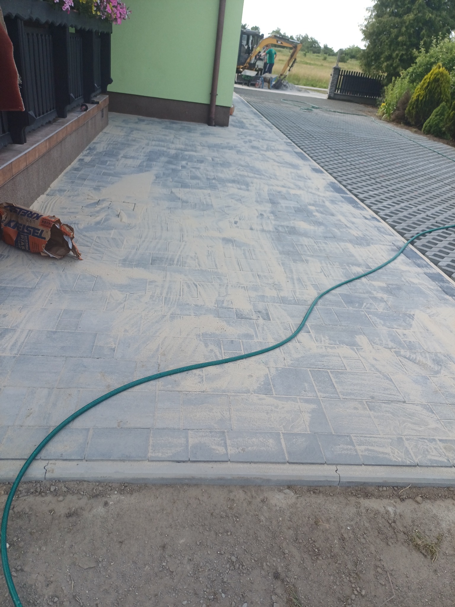 Ułożona szara kostka brukowa przed domem, częściowo zasypana piaskiem. W tle widoczny trawnik i maszyna budowlana. Na kostce leży zielony wąż ogrodowy i worek z piaskiem.
