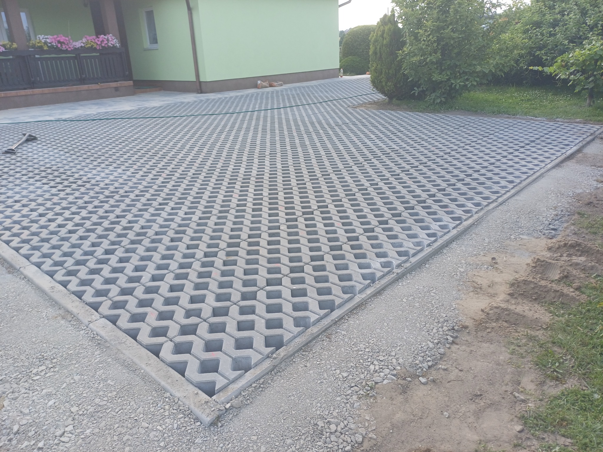 Szara ażurowa kostka ułożona na podjeździe przed domem. Widoczne obrzeża i podsypka. Częściowo widoczny budynek z zieloną elewacją i roślinność.