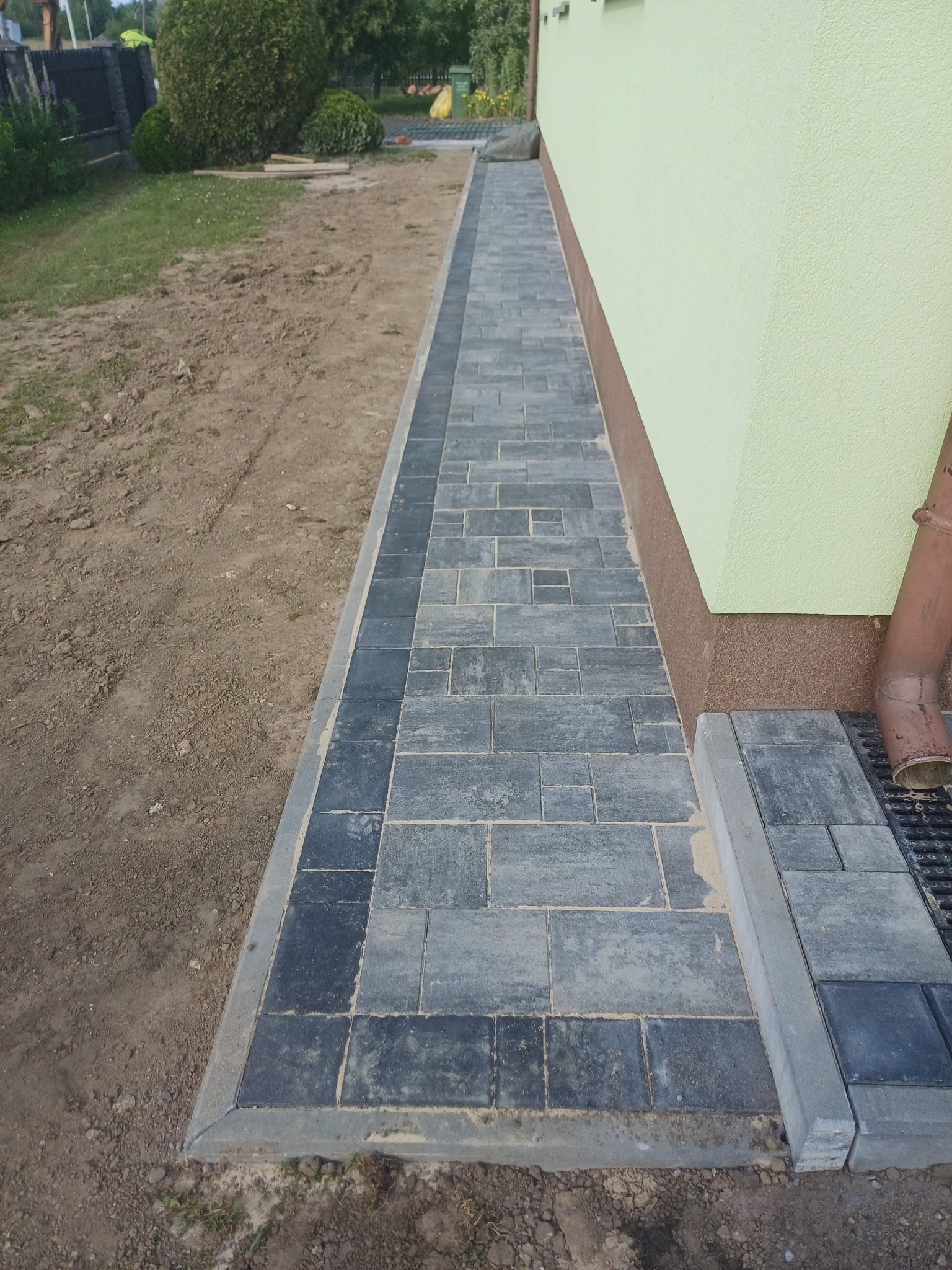 Ułożona kostka brukowa wzdłuż ściany budynku o jasnej elewacji, z widocznym odpływem liniowym. Ciemnoszara kostka w regularnym wzorze.