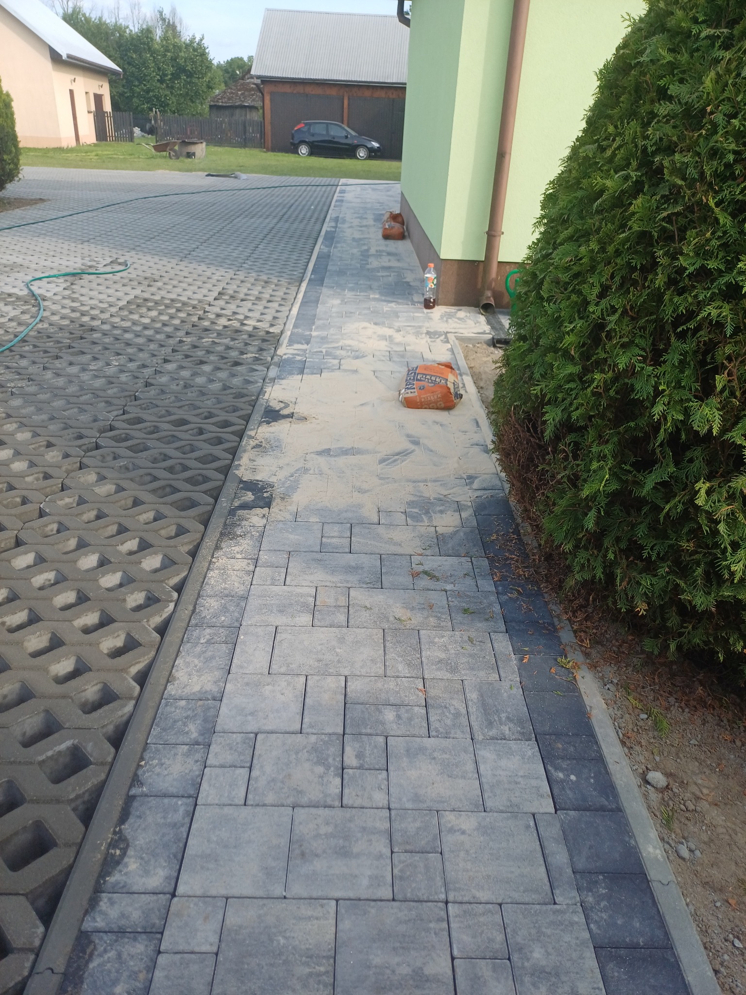 Ułożona ścieżka z szarej kostki brukowej przy zielonym budynku, obok trawnik z ażurowymi płytami. Widoczny worek z piaskiem i butelka. Praca w toku.