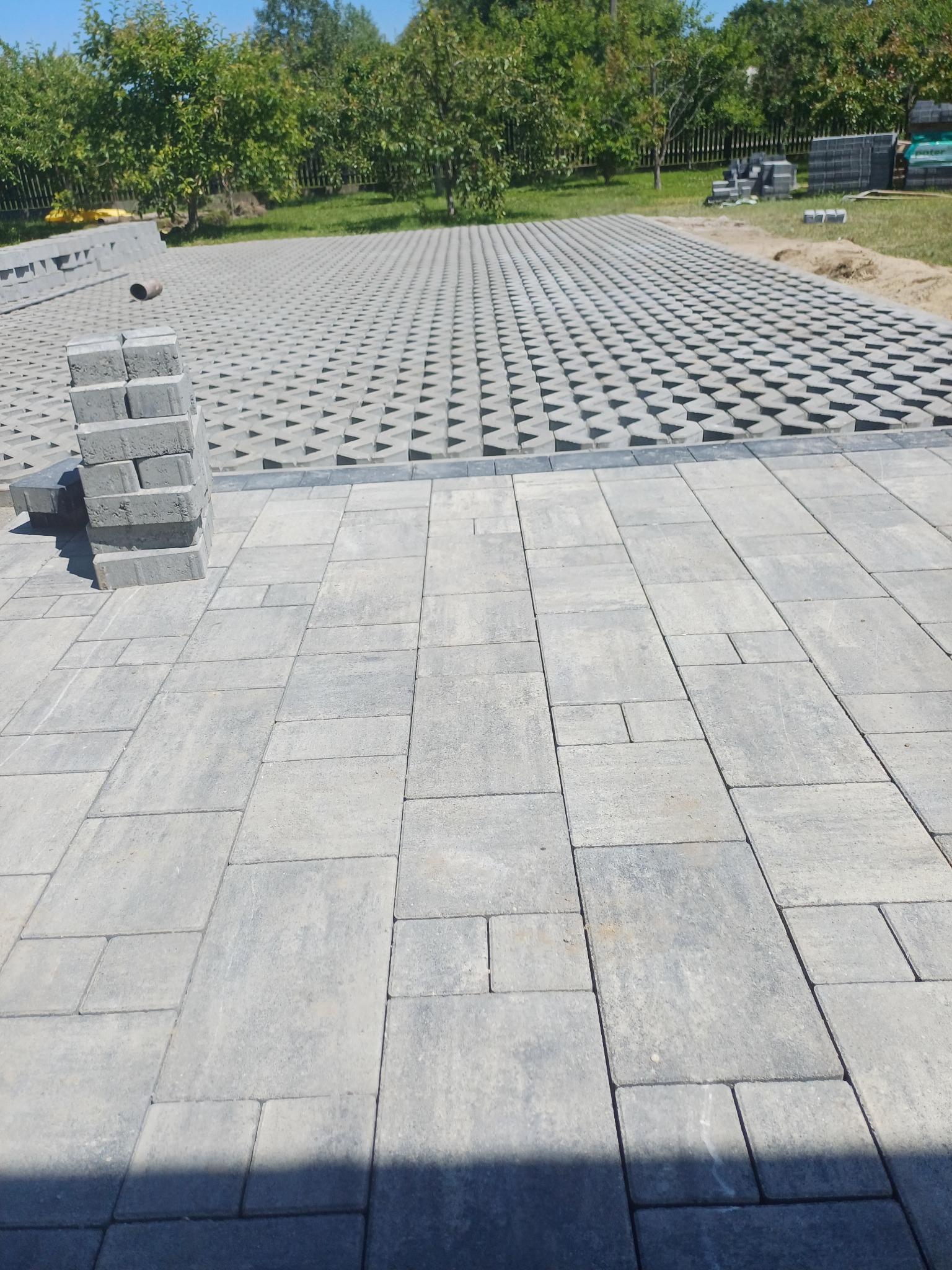 Ułożona szara kostka brukowa na podwórku, z widocznymi stertami kostki i częściowo wykończoną nawierzchnią z ażurowych płyt. Tło stanowią drzewa.