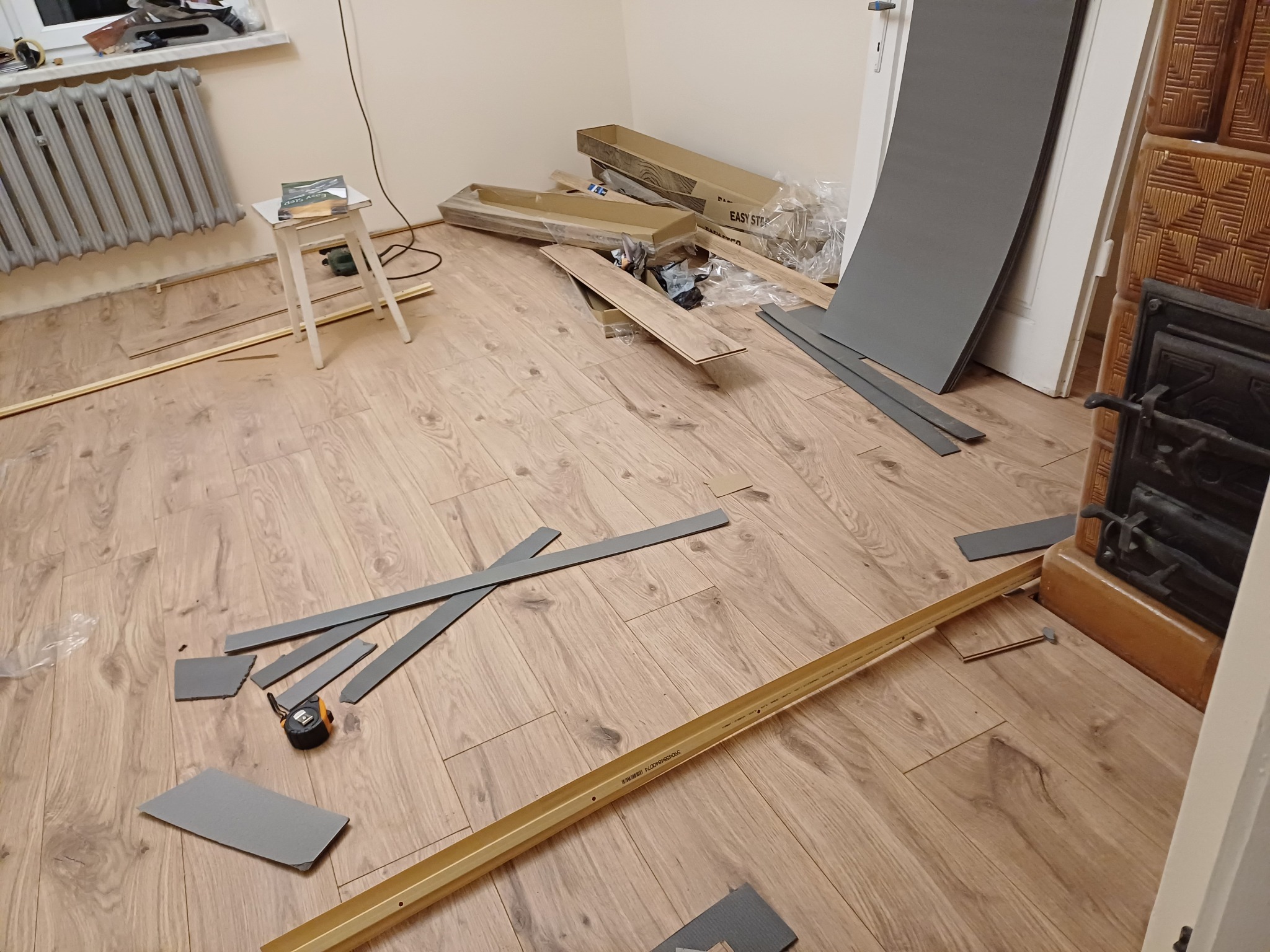 Proces układania paneli podłogowych w pokoju: rozłożone panele, listwy, narzędzia pomiarowe, podkład i opakowania. Widoczny fragment pieca kaflowego.