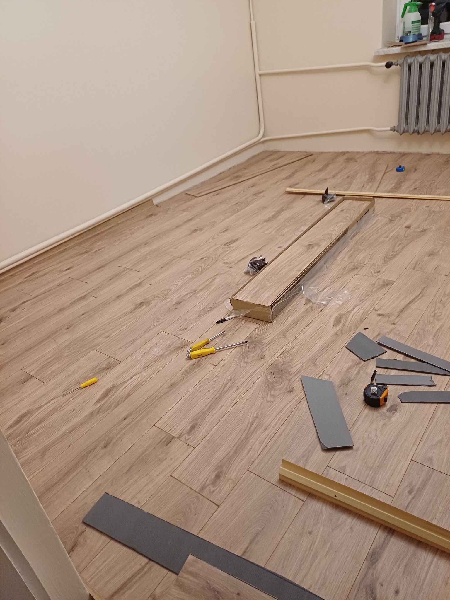 Montaż paneli podłogowych w jasnym wnętrzu. Widoczne narzędzia, listwy przypodłogowe i częściowo ułożona podłoga z paneli imitujących drewno.