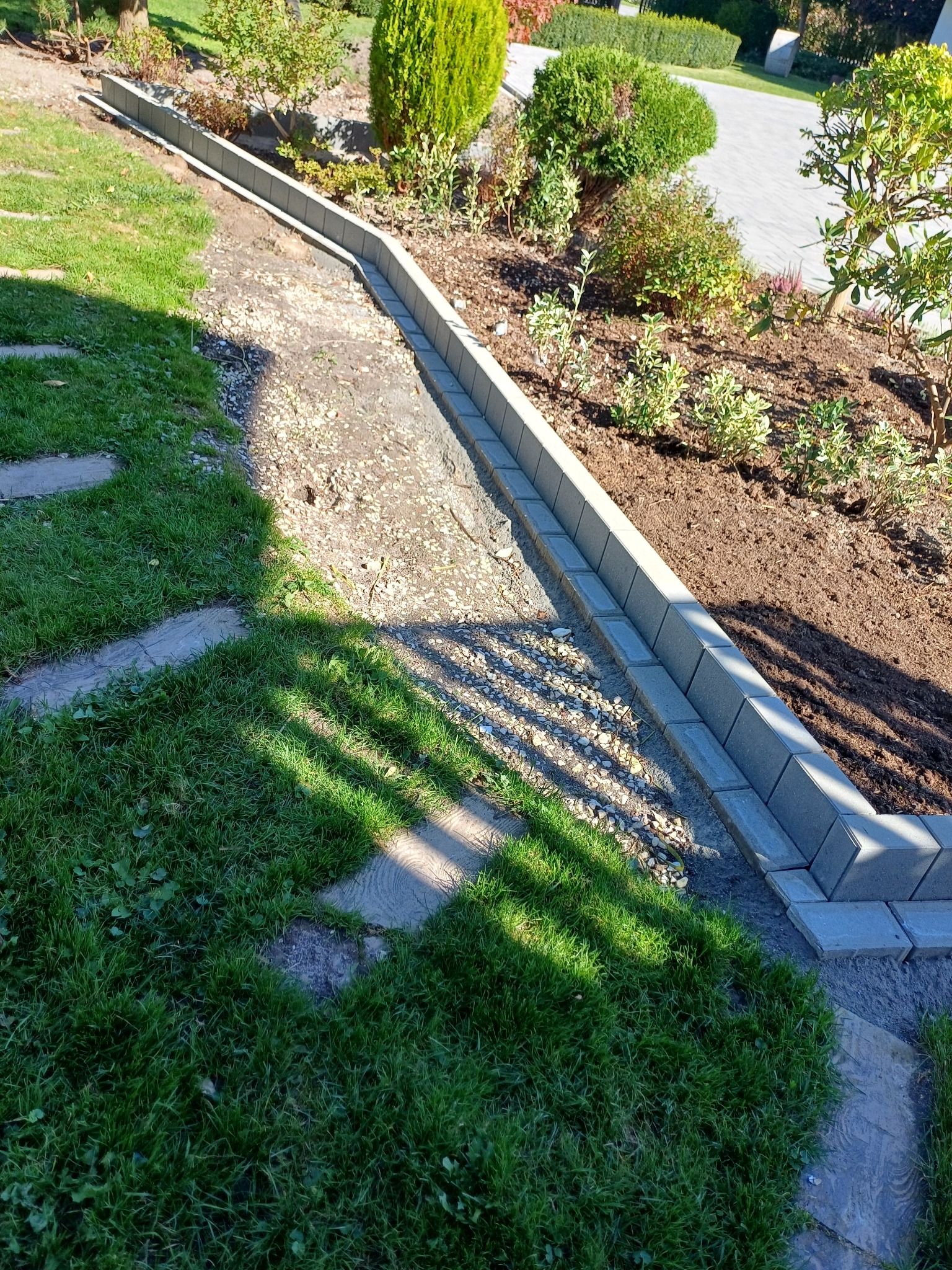 Krawężnik z kostki obrzeżający rabatę z ziemią i roślinami, widok z góry, częściowo w cieniu drzew, z elementami trawnika i ścieżki z płyt.