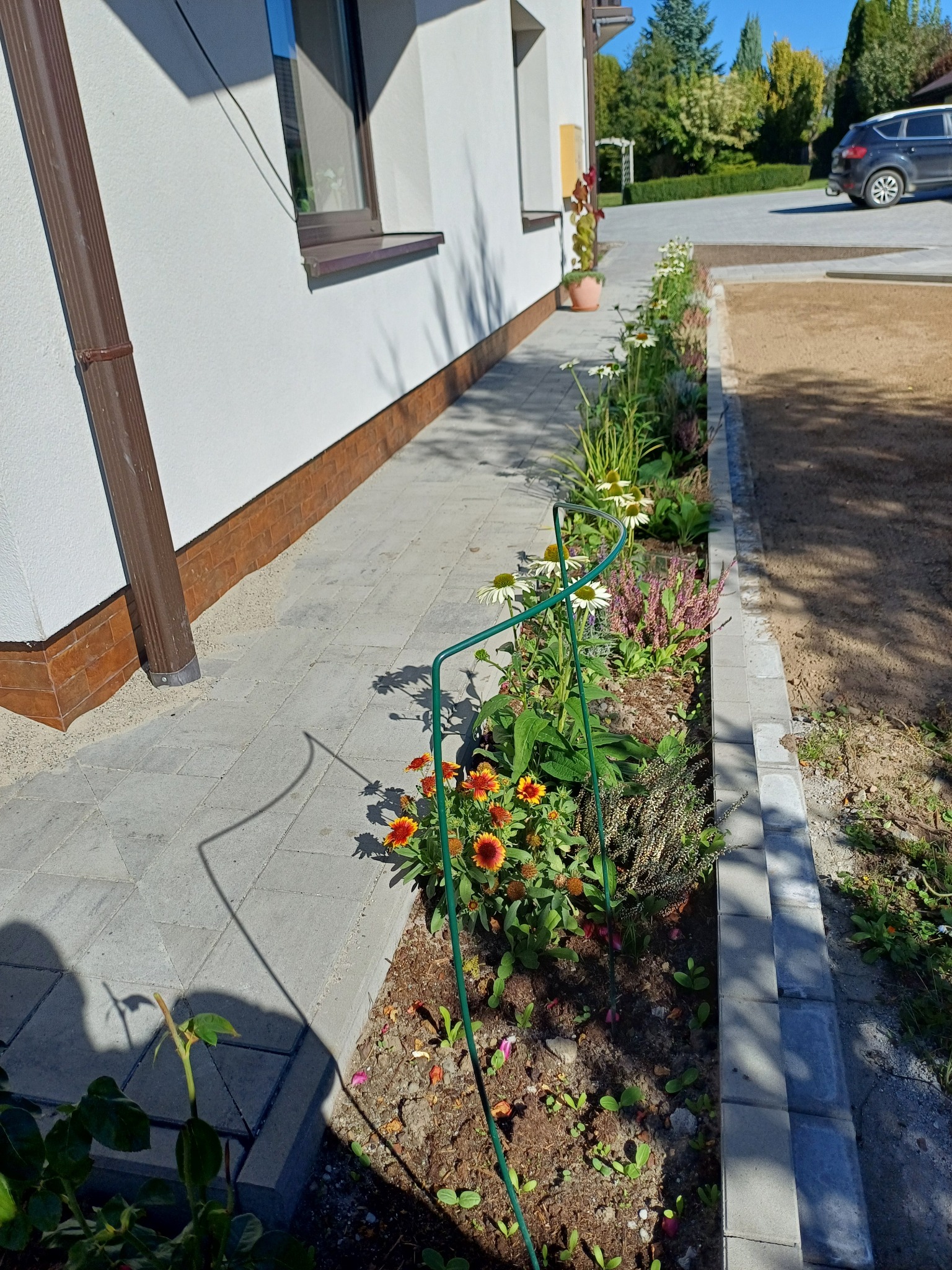 Ścieżka z kostki brukowej prowadząca wzdłuż domu, z rabatą kwiatową po prawej stronie. Widoczny fragment trawnika i zaparkowany samochód w tle.