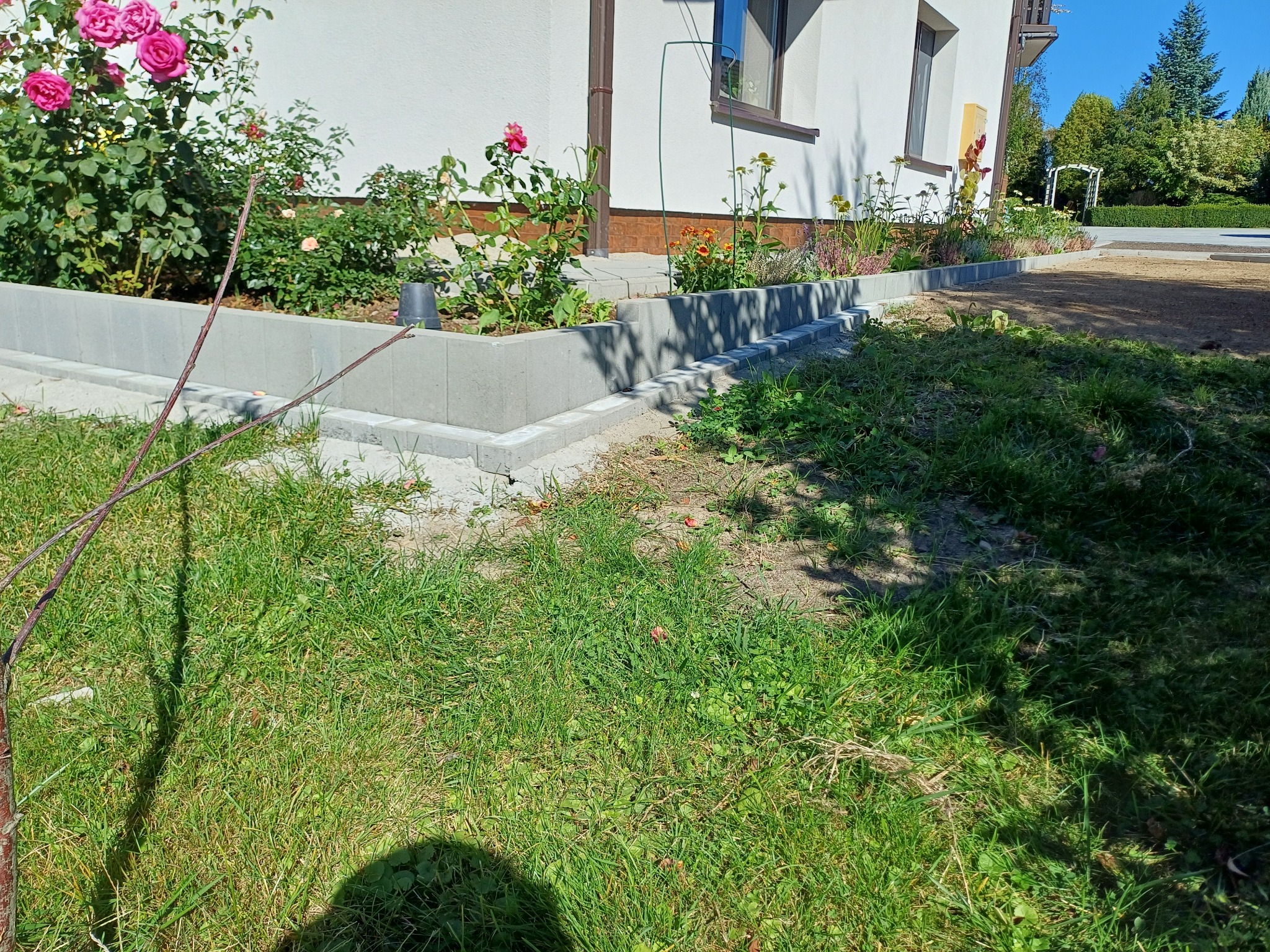 Obrzeża rabat z szarej kostki brukowej, wykończone z dbałością o detale, otaczające rabaty kwiatowe przy domu w Pruchnik. Widoczne świeże nasadzenia.