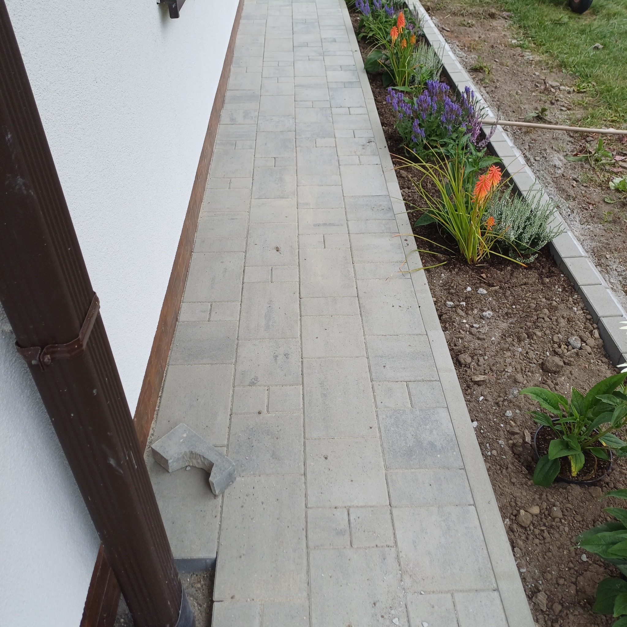 Ścieżka z jasnoszarej kostki brukowej prowadząca wzdłuż białej ściany budynku, z rabatą kwiatową po prawej stronie. Fragment kostki leży obok rynny.
