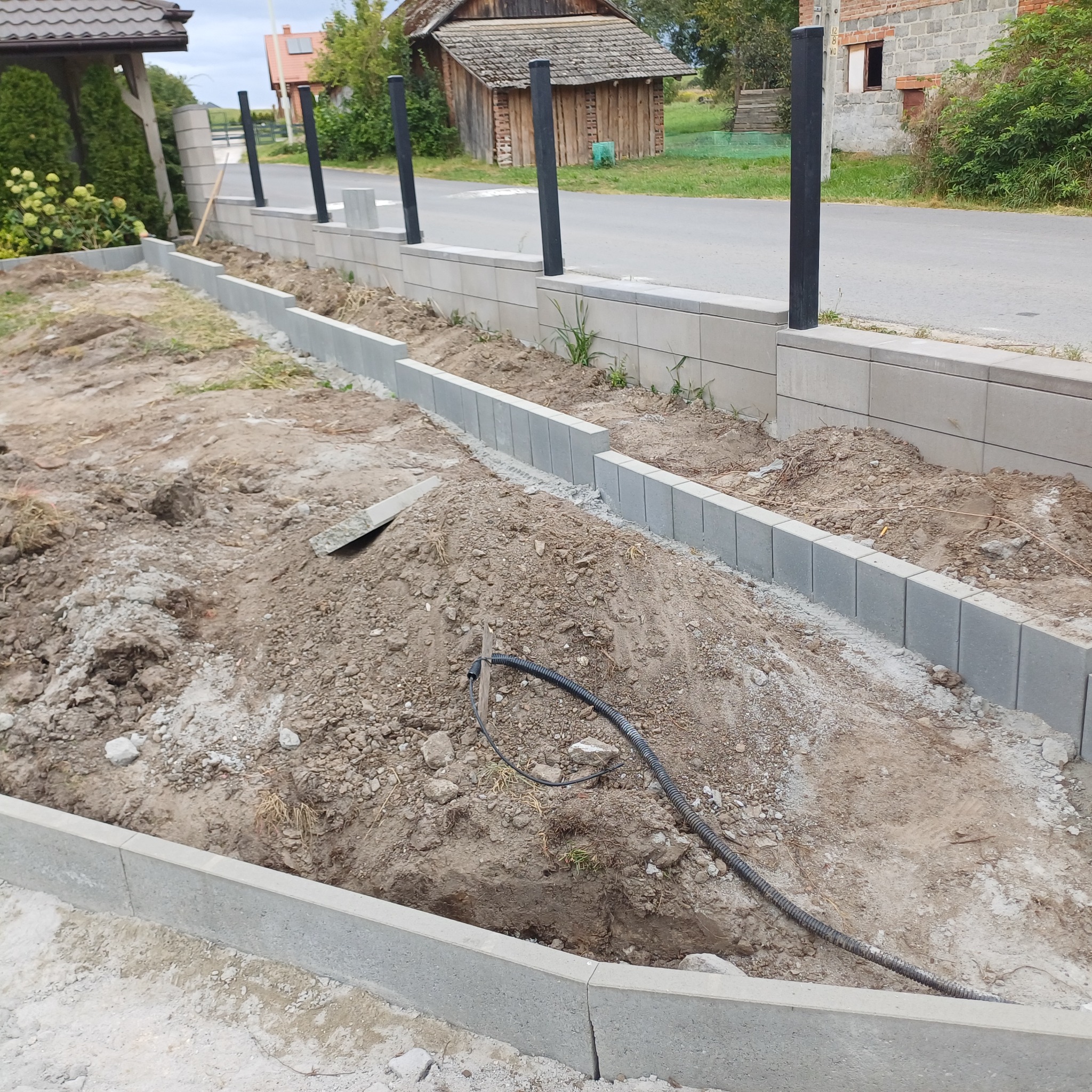 Wyrównany teren z obrzeżami z szarej kostki betonowej, przygotowany pod dalsze prace brukarskie. W tle widoczny fragment ogrodzenia i budynki.