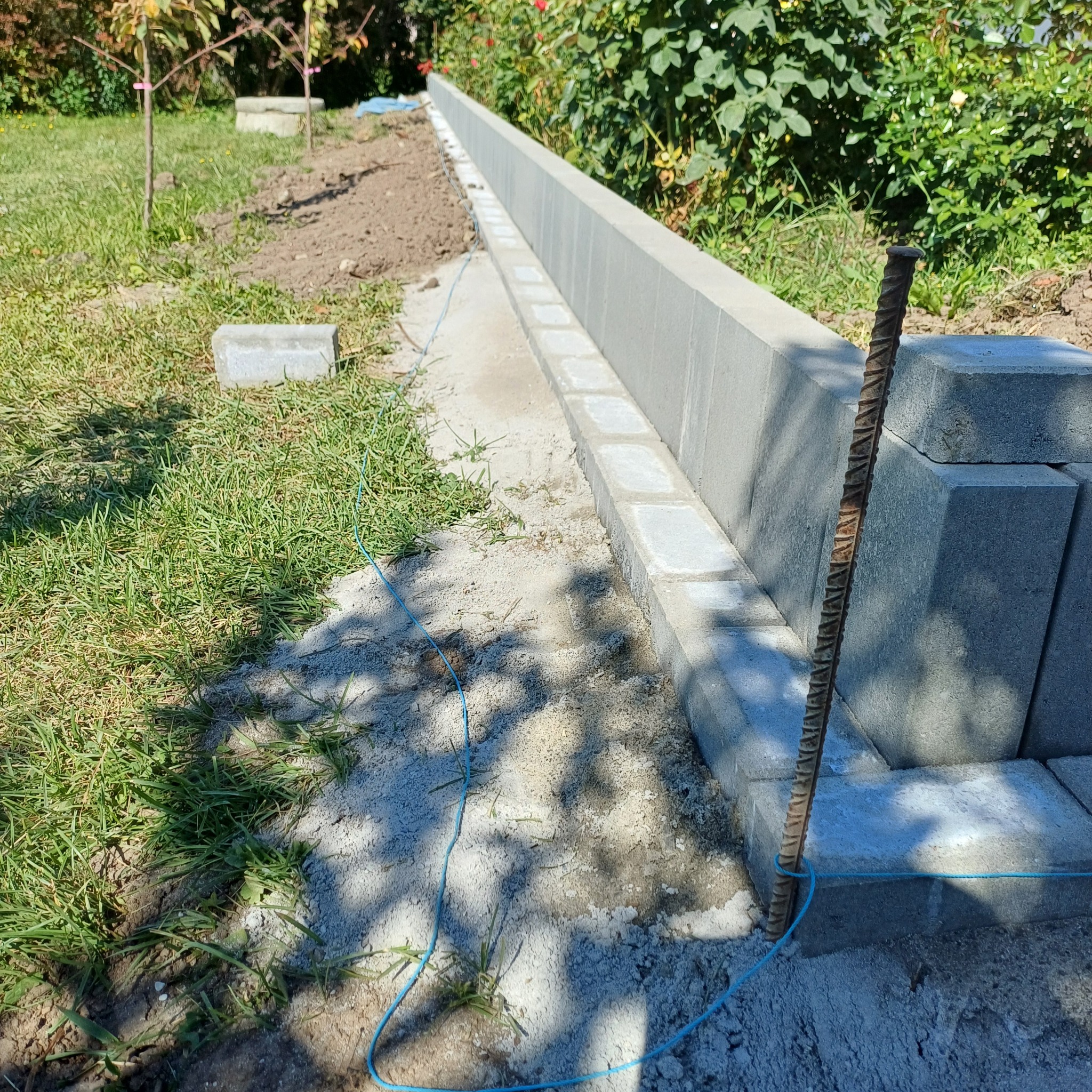 Wyrównany teren pod budowę murku z betonowych bloczków, widoczne sznurki wyznaczające linię, fragment trawnika i roślinność w tle. Prace ziemne w toku.