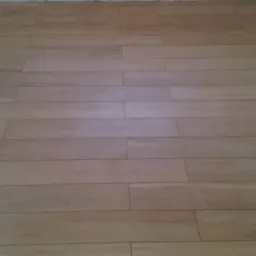 Ułożona podłoga z długich, prostokątnych płytek imitujących drewno, w odcieniach beżu i brązu. Płytki ułożone poziomo, widoczne fugi. Fragment ściany w tle.