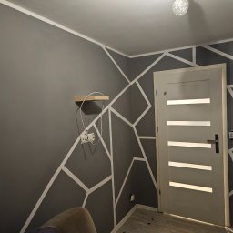 GOODesigne - Szara ściana z geometrycznym wzorem z białych pasków, drewniana półka i szara, nowoczesna, wewnętrzna stolarka drzwiowa z poziomymi przeszkleniami.