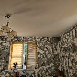 GOODesigne - Pokój po szpachlowaniu dekoracyjnym: ściany w odcieniach szarości i bieli z wyraźną fakturą, okno z żaluzjami, sufit biały z lampą. Widoczne ślady pracy.