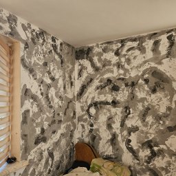 GOODesigne - Ściana z dekoracyjną fakturą w odcieniach szarości i bieli, widok z bliska na wykończone wnętrze z oknem z żaluzjami poziomymi i widocznym grzejnikiem.