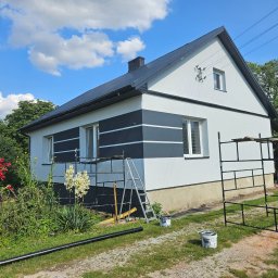 GOODesigne - Dom z pomalowaną fasadą w biało-czarne pasy, otoczony zielenią. Widoczne rusztowania, drabina i akcesoria malarskie, sugerujące prace wykończeniowe na zewnątrz budynku.