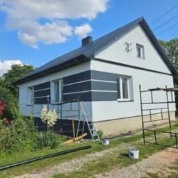 Dom z pomalowaną fasadą w biało-czarne pasy, otoczony zielenią. Widoczne rusztowania, drabina i akcesoria malarskie, sugerujące prace wykończeniowe na zewnątrz budynku.