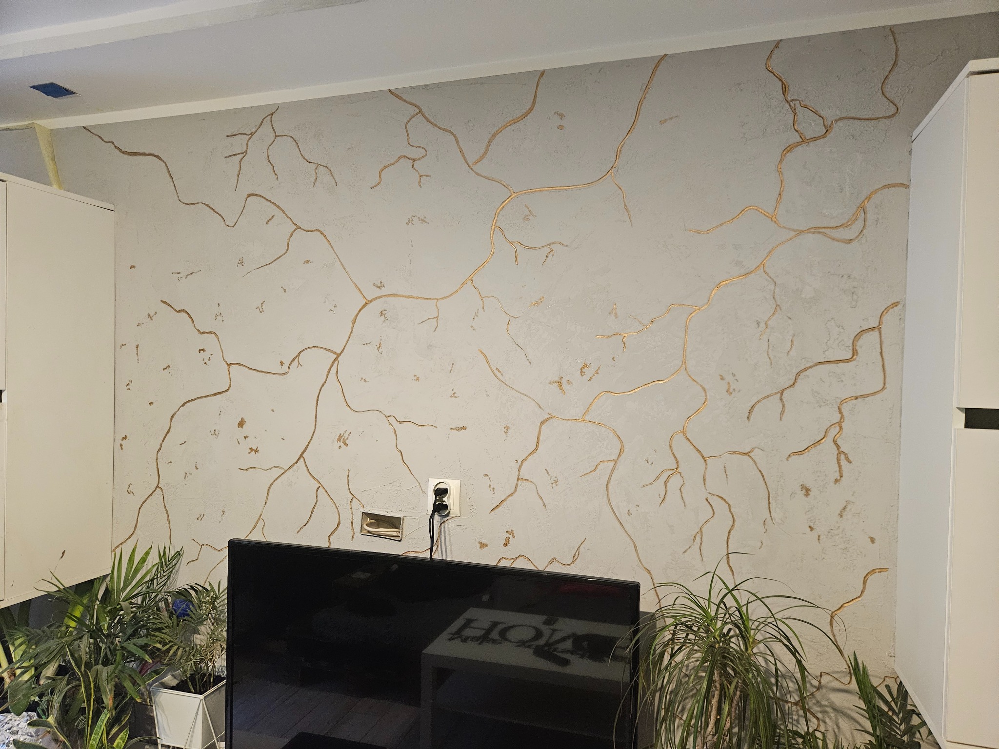 Ściana z dekoracyjnym tynkiem w odcieniach szarości, z nieregularnymi, złotymi wzorami przypominającymi pęknięcia. Widoczny fragment telewizora i rośliny doniczkowe.