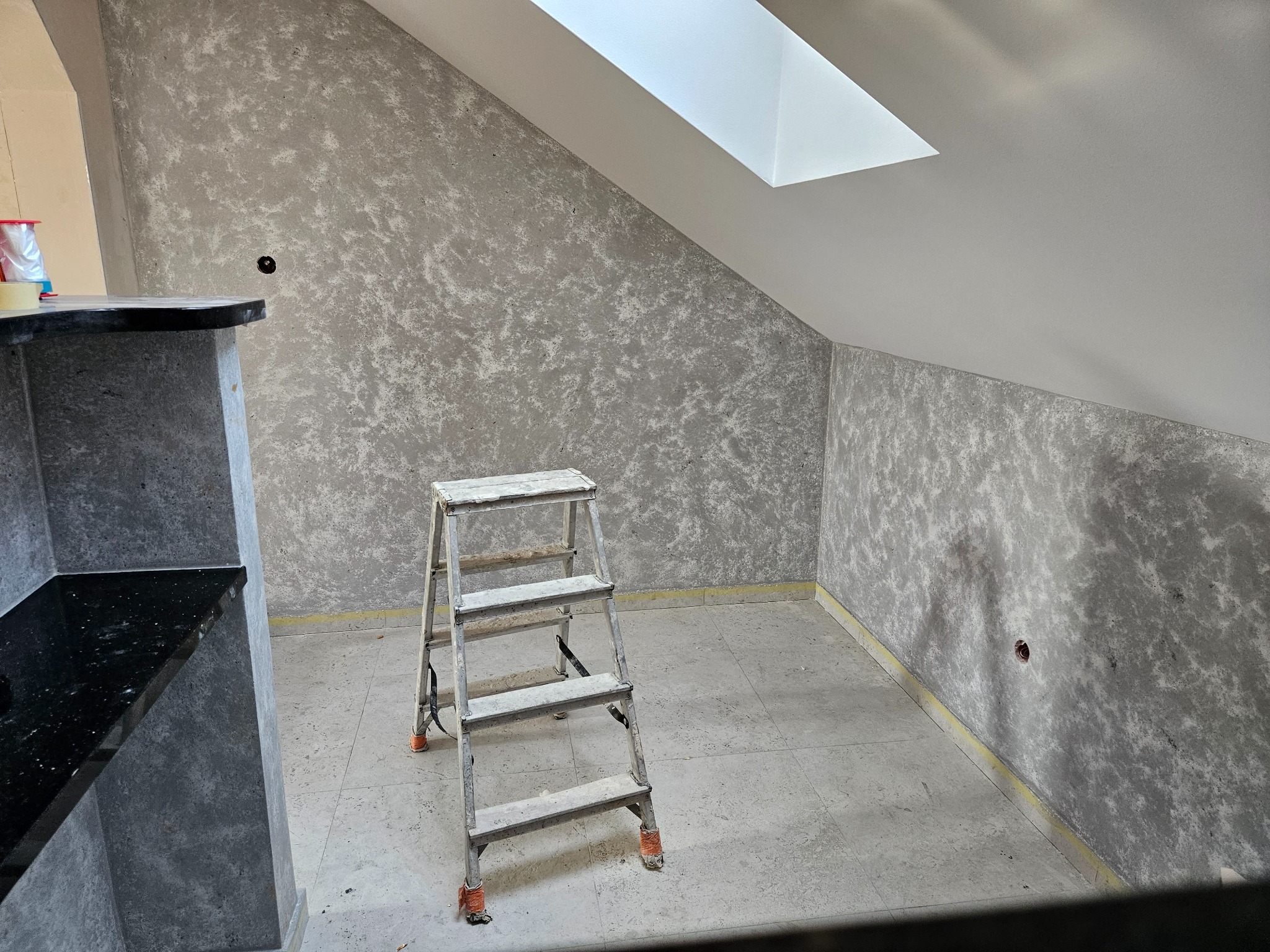 Wykończone ściany w szarym odcieniu z efektem dekoracyjnym, drabina aluminiowa w pomieszczeniu na poddaszu z oknem dachowym. Widoczne gniazdka elektryczne.
