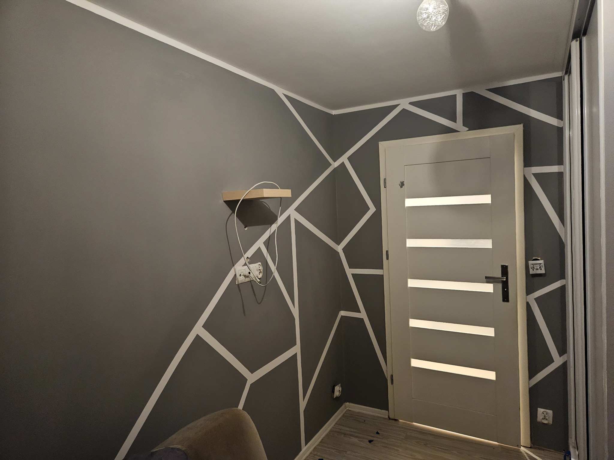 Szara ściana z geometrycznym wzorem z białych pasków, drewniana półka i szara, nowoczesna, wewnętrzna stolarka drzwiowa z poziomymi przeszkleniami.