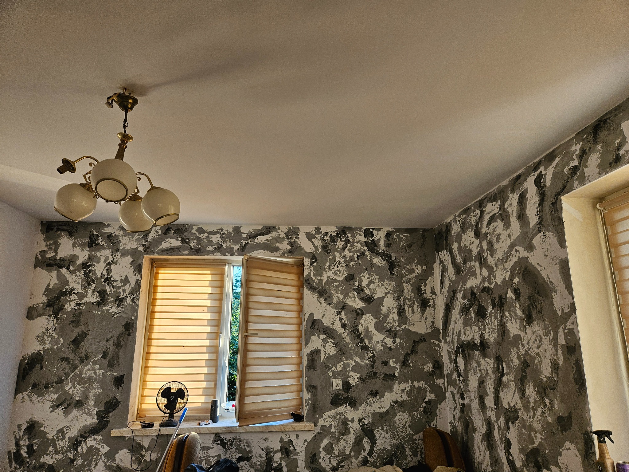 Pokój po szpachlowaniu dekoracyjnym: ściany w odcieniach szarości i bieli z wyraźną fakturą, okno z żaluzjami, sufit biały z lampą. Widoczne ślady pracy.