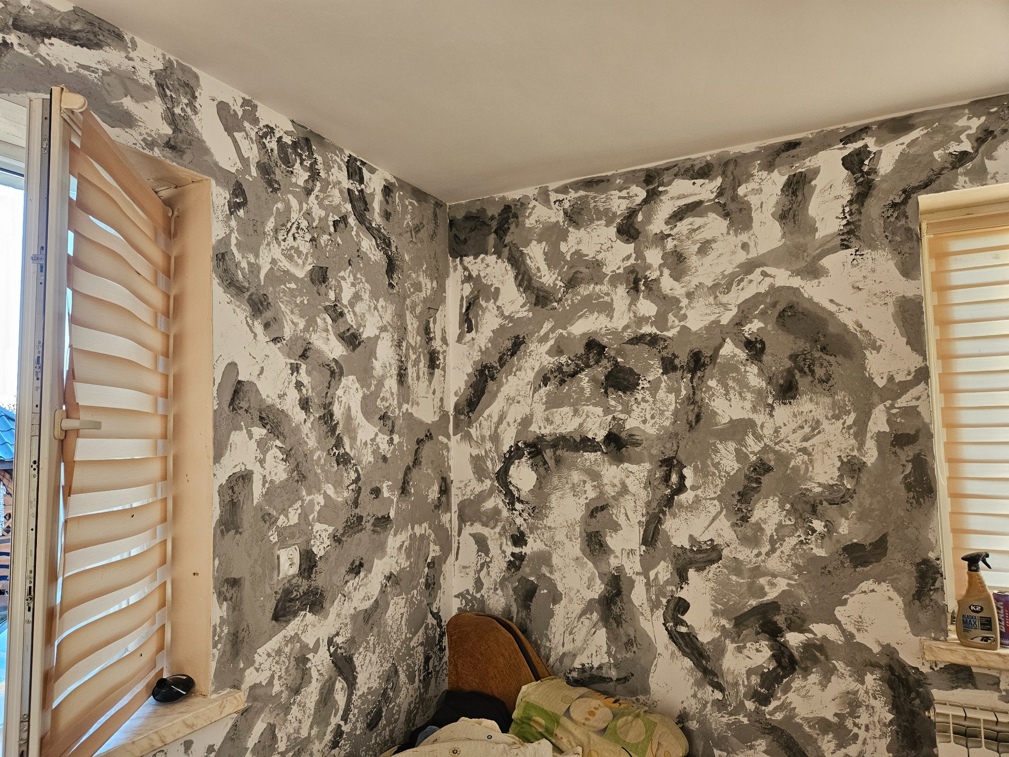 Ściana z dekoracyjną fakturą w odcieniach szarości i bieli, widok z bliska na wykończone wnętrze z oknem z żaluzjami poziomymi i widocznym grzejnikiem.