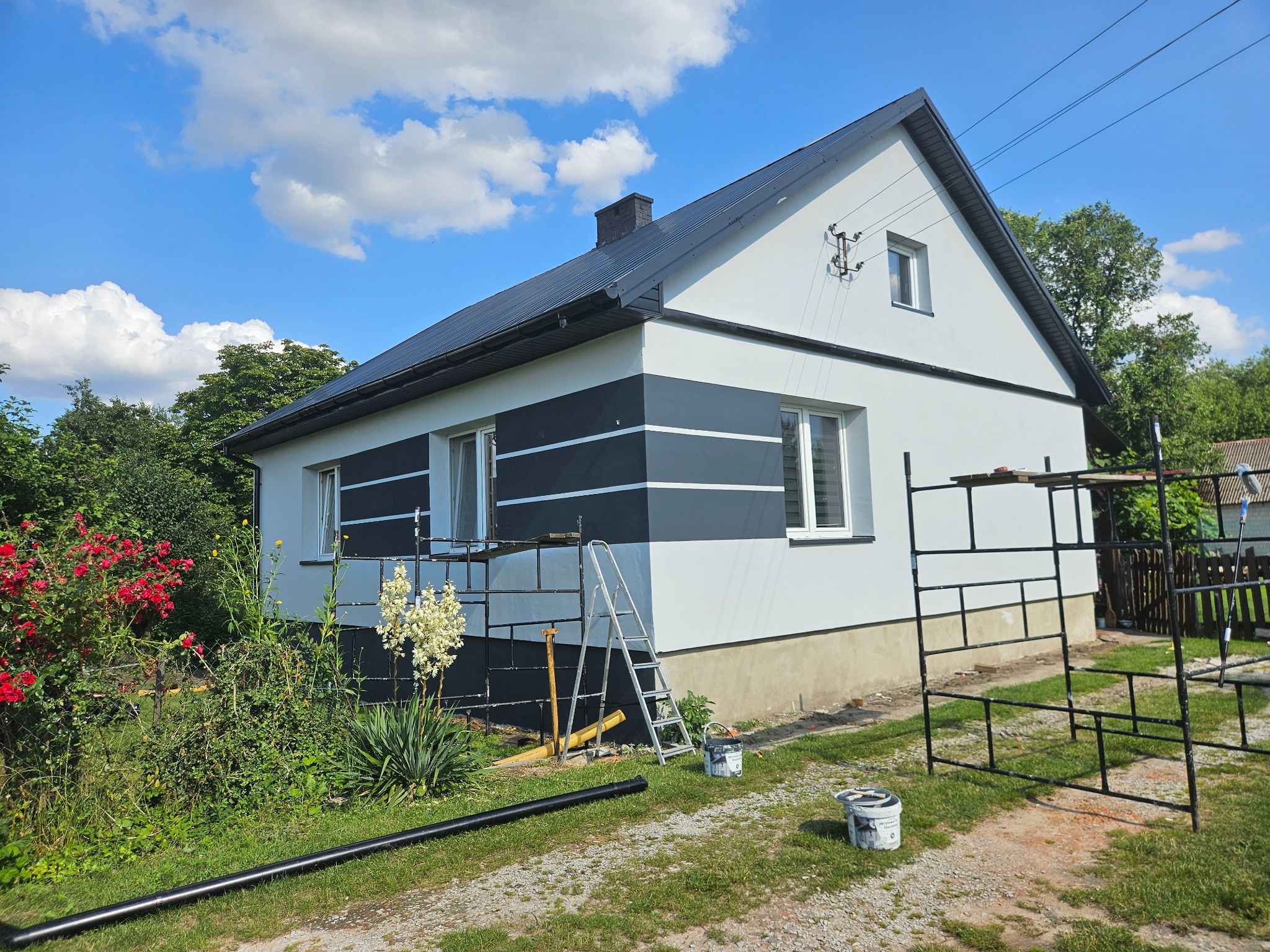 Dom z pomalowaną fasadą w biało-czarne pasy, otoczony zielenią. Widoczne rusztowania, drabina i akcesoria malarskie, sugerujące prace wykończeniowe na zewnątrz budynku.