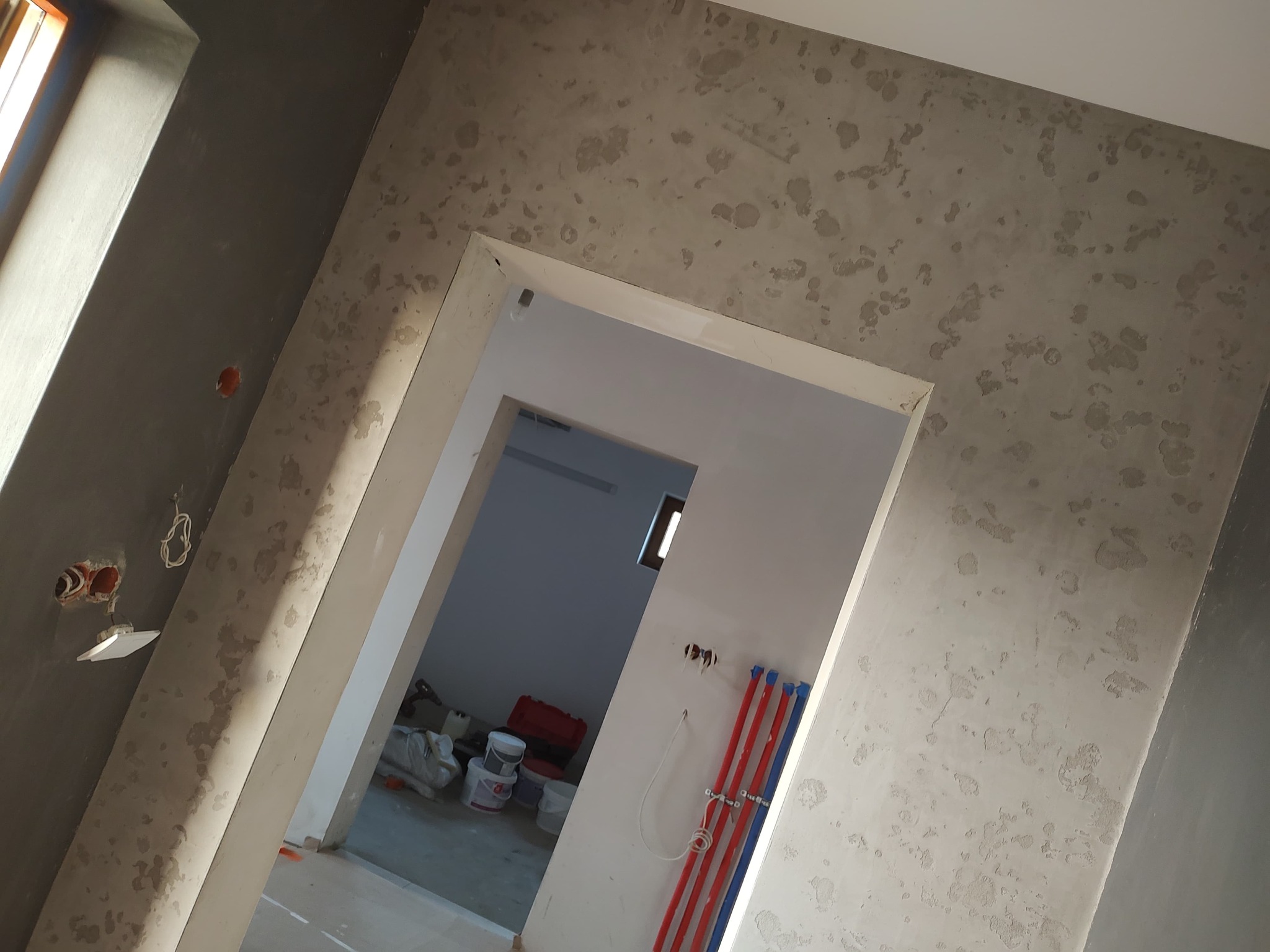 Ściana z efektem dekoracyjnym w odcieniach szarości, widok przez białe futryny drzwiowe. W tle widoczne przygotowane materiały budowlane i instalacje.