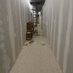 Merob Sp.z o.o. - Korytarz w trakcie wykańczania: ściany pokryte gładzią, kartony z materiałami budowlanymi, drabina w głębi. Widok w perspektywie, surowy beton na suficie.