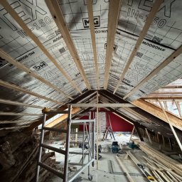 Merob Sp.z o.o. - Poddasze w trakcie prac wykończeniowych: widoczne krokwie, izolacja TEX-IN-150, rusztowanie i narzędzia. Czerwona ściana w tle. Widać etap prac.