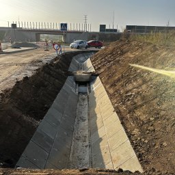 Merob Sp.z o.o. - Betonowy rów odwadniający w wykopie, z widocznym mostem w tle i znakami drogowymi. Prace ziemne w toku, teren budowy.