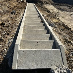 Merob Sp.z o.o. - Długie, betonowe schody zewnętrzne prowadzą w górę zbocza. Ułożone z szarych płyt, z widocznym butem w rogu kadru. Tło to ziemia i zbocze.