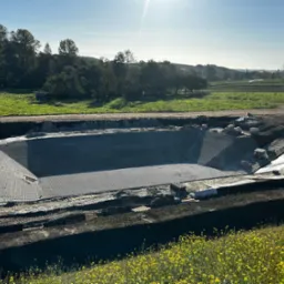 Wyłożony siatką drenażową wykop pod zbiornik wodny na tle zielonego krajobrazu z żółtymi kwiatami. W tle widoczne drzewa i pola uprawne.