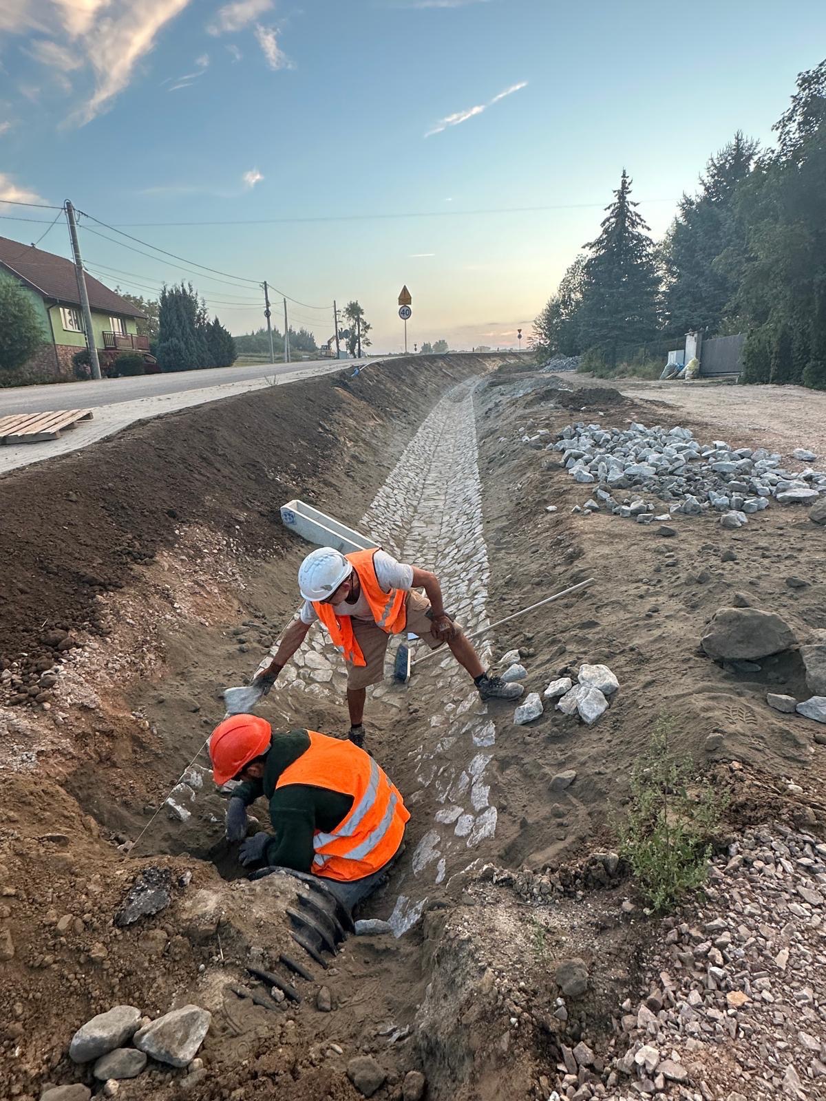 Dwóch robotników w kamizelkach układa kamienną nawierzchnię w rowie wzdłuż drogi. Praca w toku, widoczne narzędzia i materiały budowlane. Krajobraz wiejski w tle.