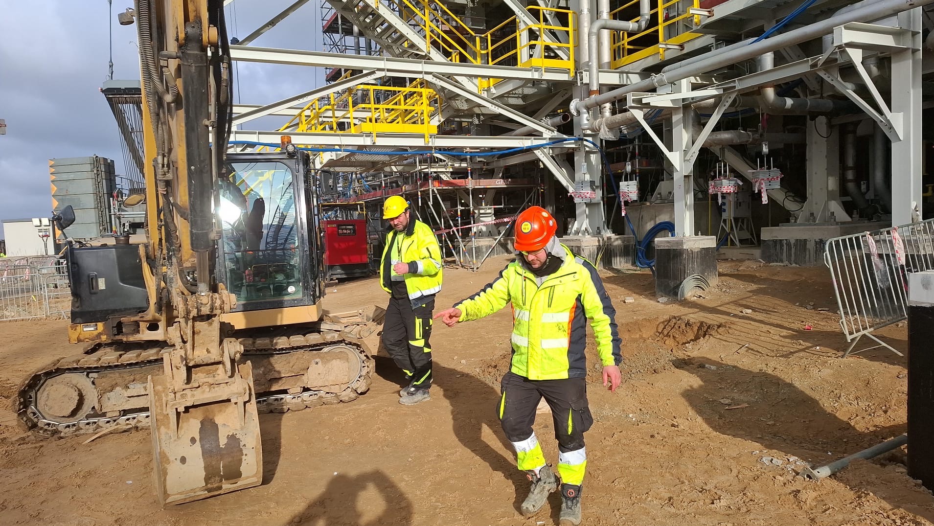 Dwóch pracowników w kaskach i odblaskowych kurtkach obok koparki na placu budowy z metalową konstrukcją w tle, jeden wskazuje palcem.