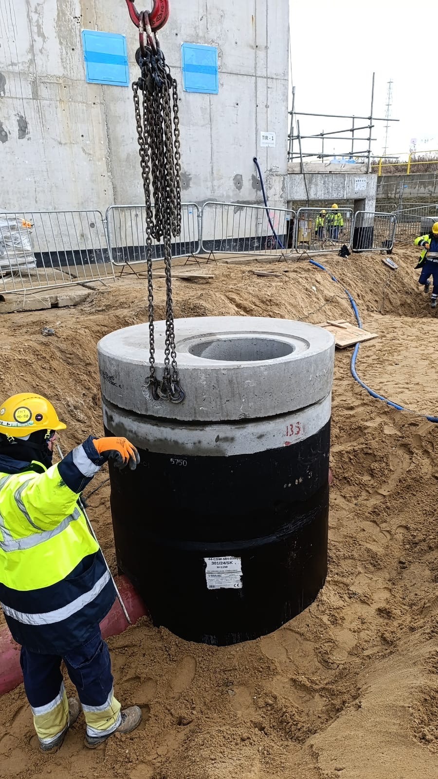 Montaż betonowej studni kanalizacyjnej na placu budowy. Robotnik w kasku i kamizelce odblaskowej nadzoruje proces. Elementy studni zawieszone na łańcuchach.