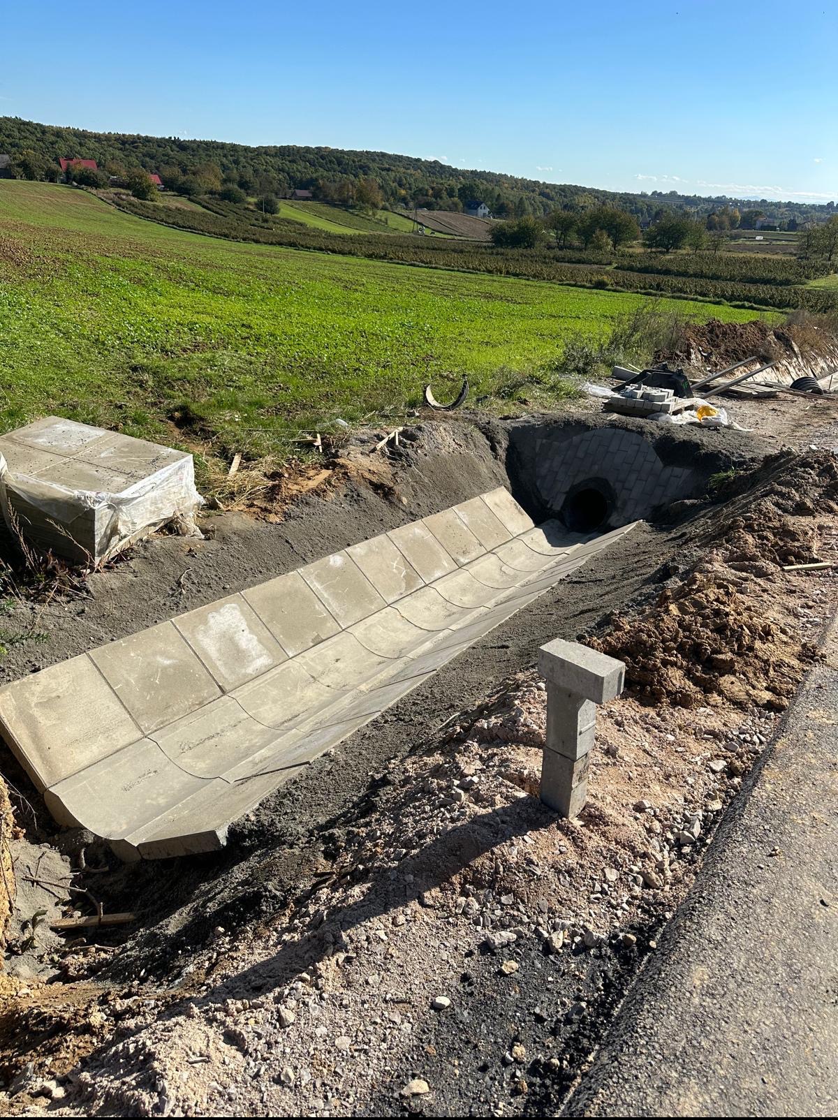 Betonowy kanał odwadniający, częściowo ułożony w ziemi, z widokiem na krajobraz wiejski w tle, w słoneczny dzień. Elementy betonowe ułożone obok.
