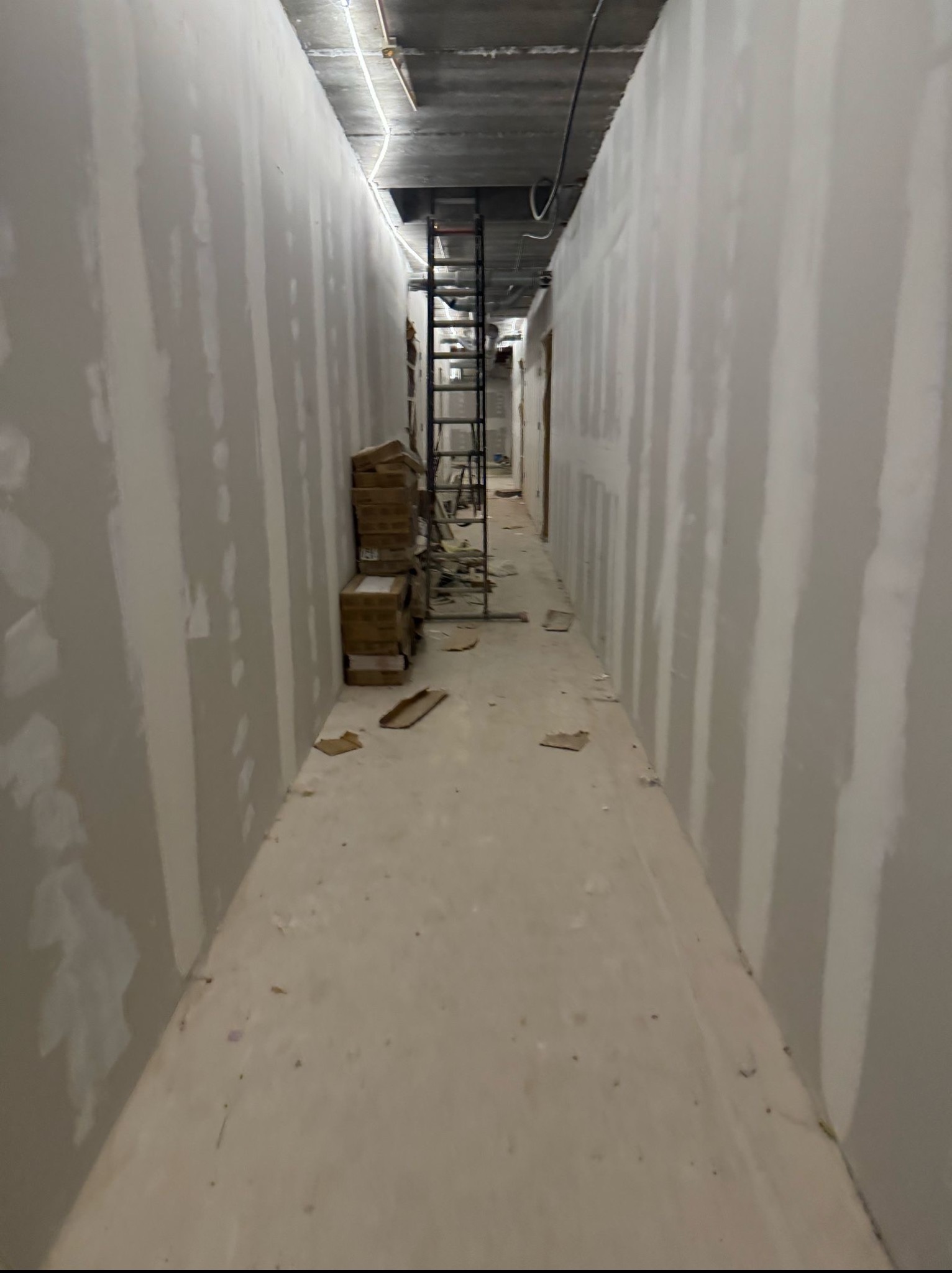 Korytarz w trakcie wykańczania: ściany pokryte gładzią, kartony z materiałami budowlanymi, drabina w głębi. Widok w perspektywie, surowy beton na suficie.