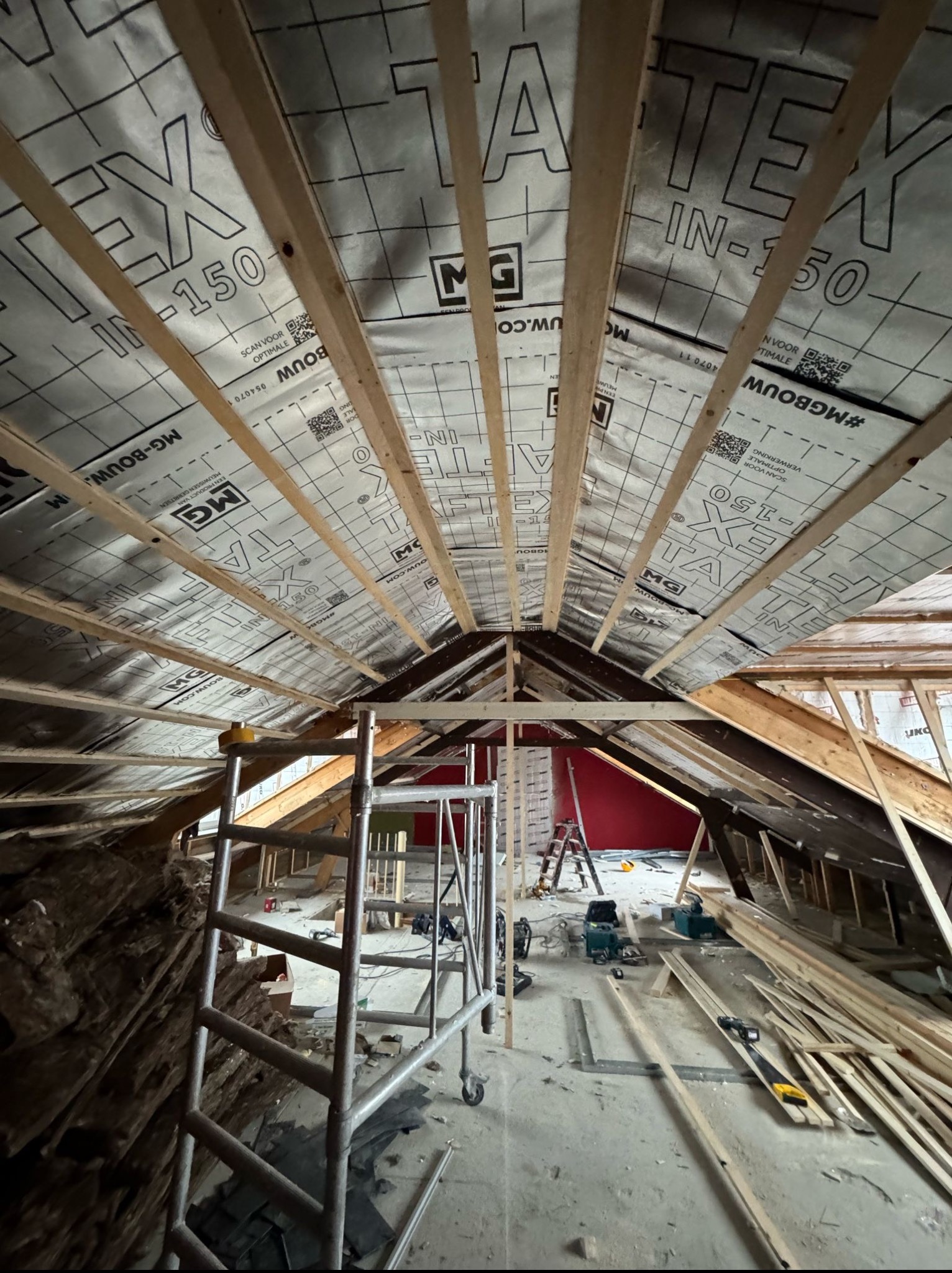 Poddasze w trakcie prac wykończeniowych: widoczne krokwie, izolacja TEX-IN-150, rusztowanie i narzędzia. Czerwona ściana w tle. Widać etap prac.