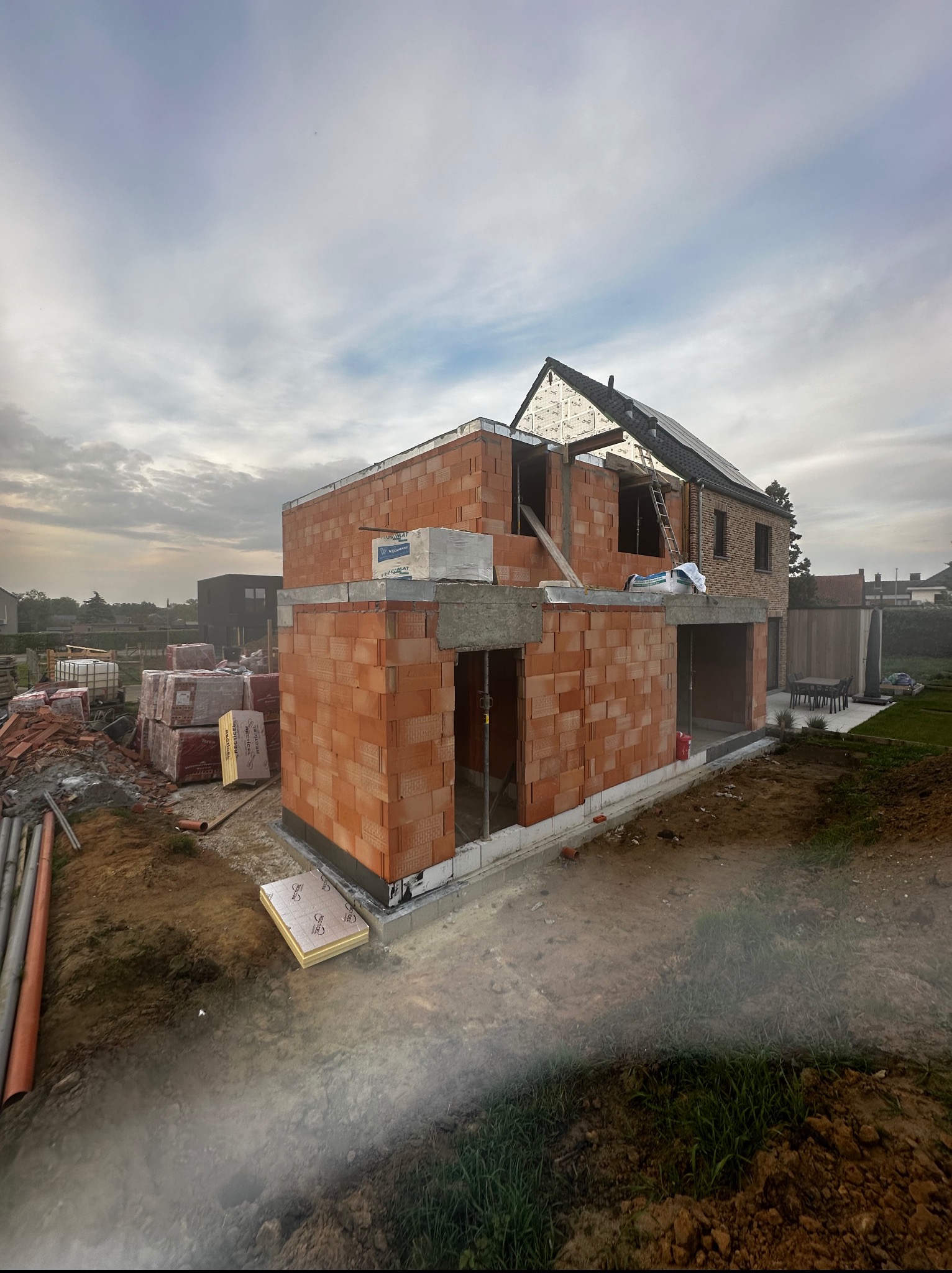 Budowa domu jednorodzinnego w trakcie wznoszenia ścian z czerwonej cegły. Częściowo widoczny dach i otoczenie budowy.