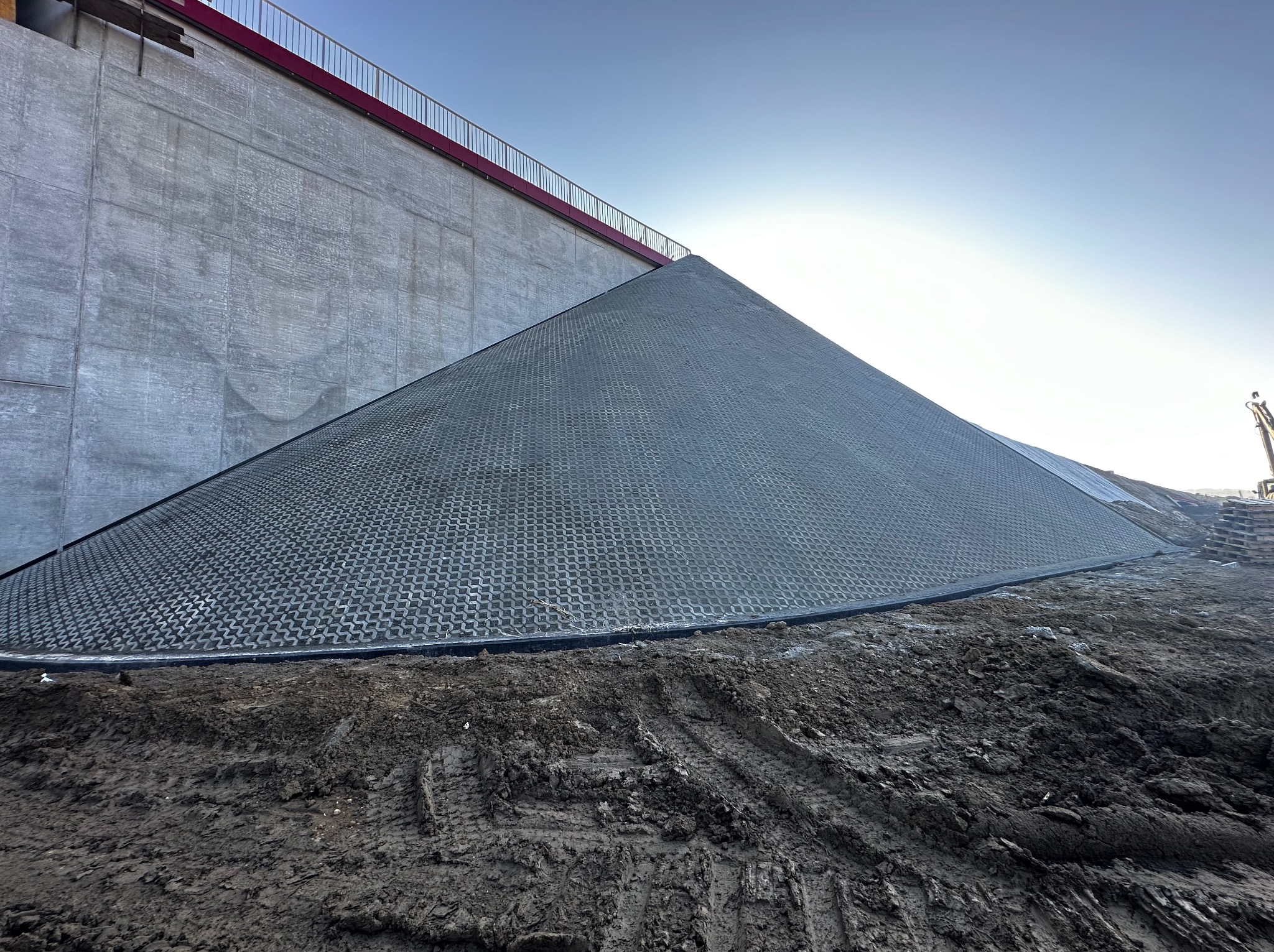 Skarpa zabezpieczona geokratą na tle betonowej ściany z czerwoną barierką. Ziemia w dole, jasne niebo w górze. Widoczne ślady opon.