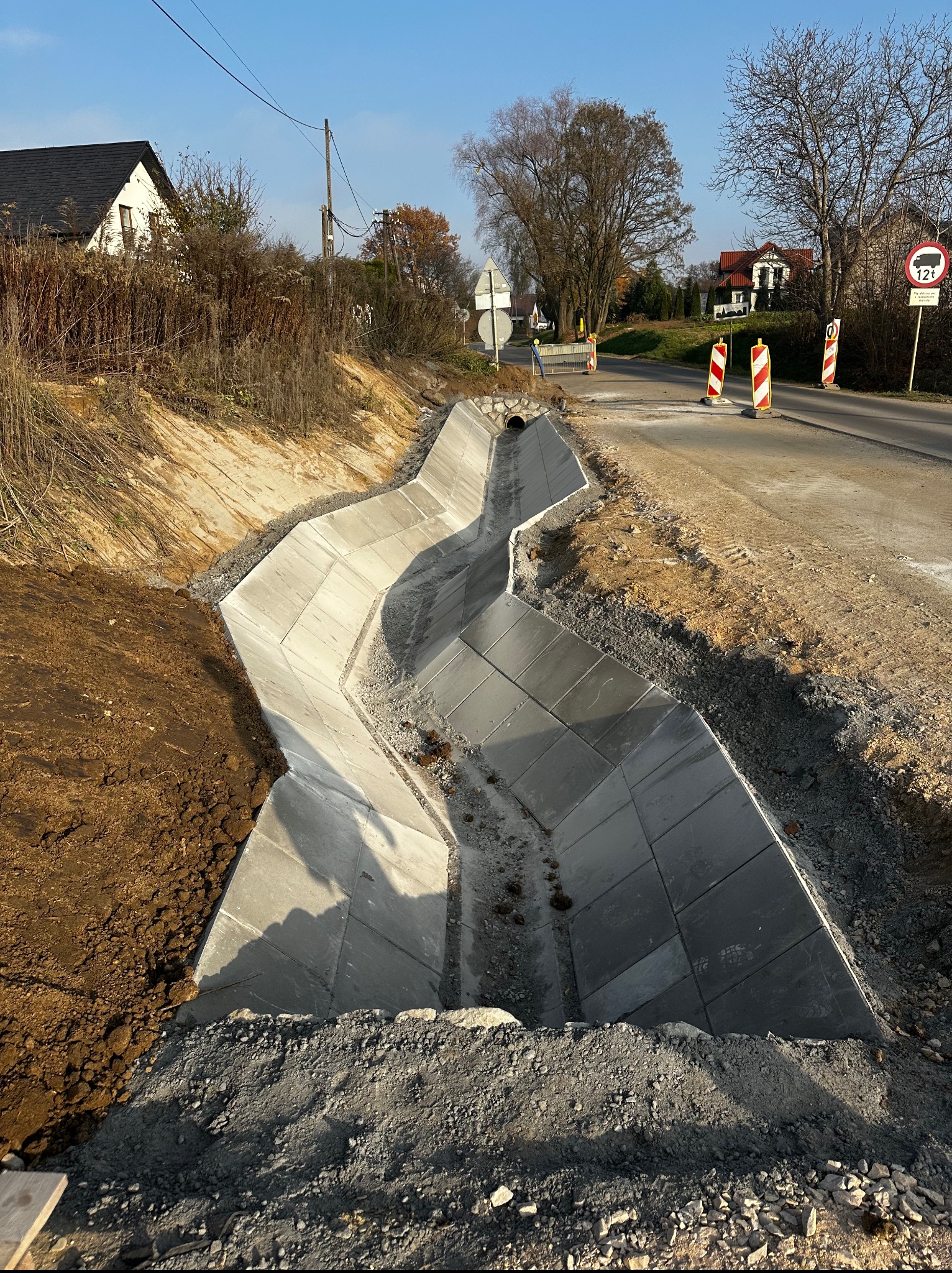 Betonowy rów melioracyjny wyłożony płytami, widoczny z góry, przylegający do asfaltowej drogi z ograniczeniem tonażu, w tle domy i drzewa.