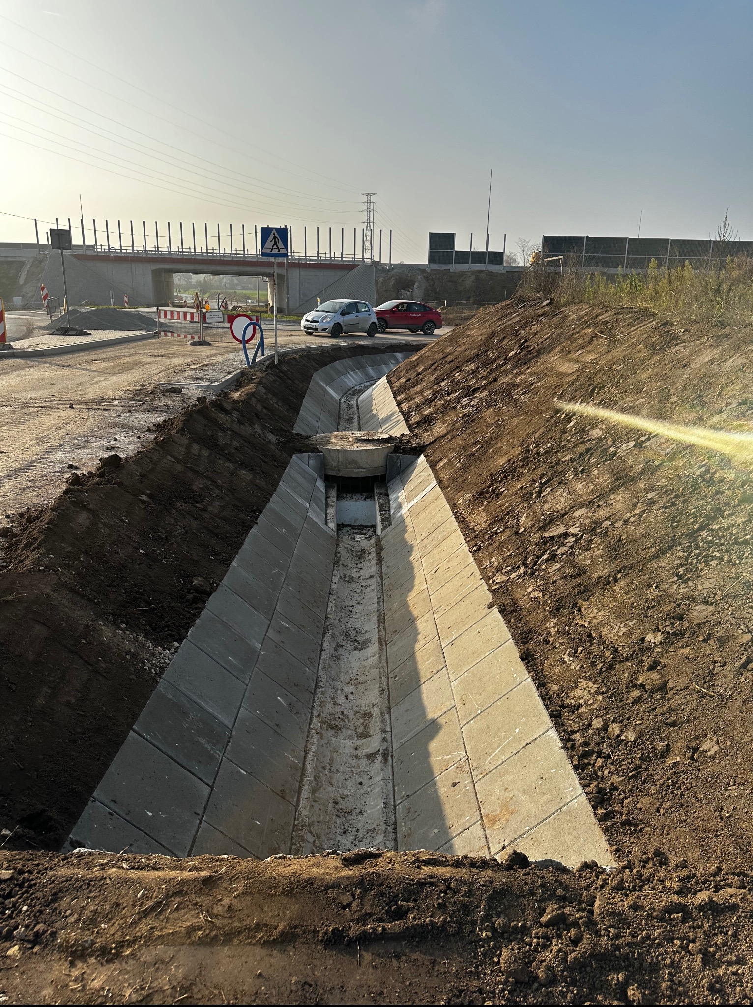 Betonowy rów odwadniający w wykopie, z widocznym mostem w tle i znakami drogowymi. Prace ziemne w toku, teren budowy.