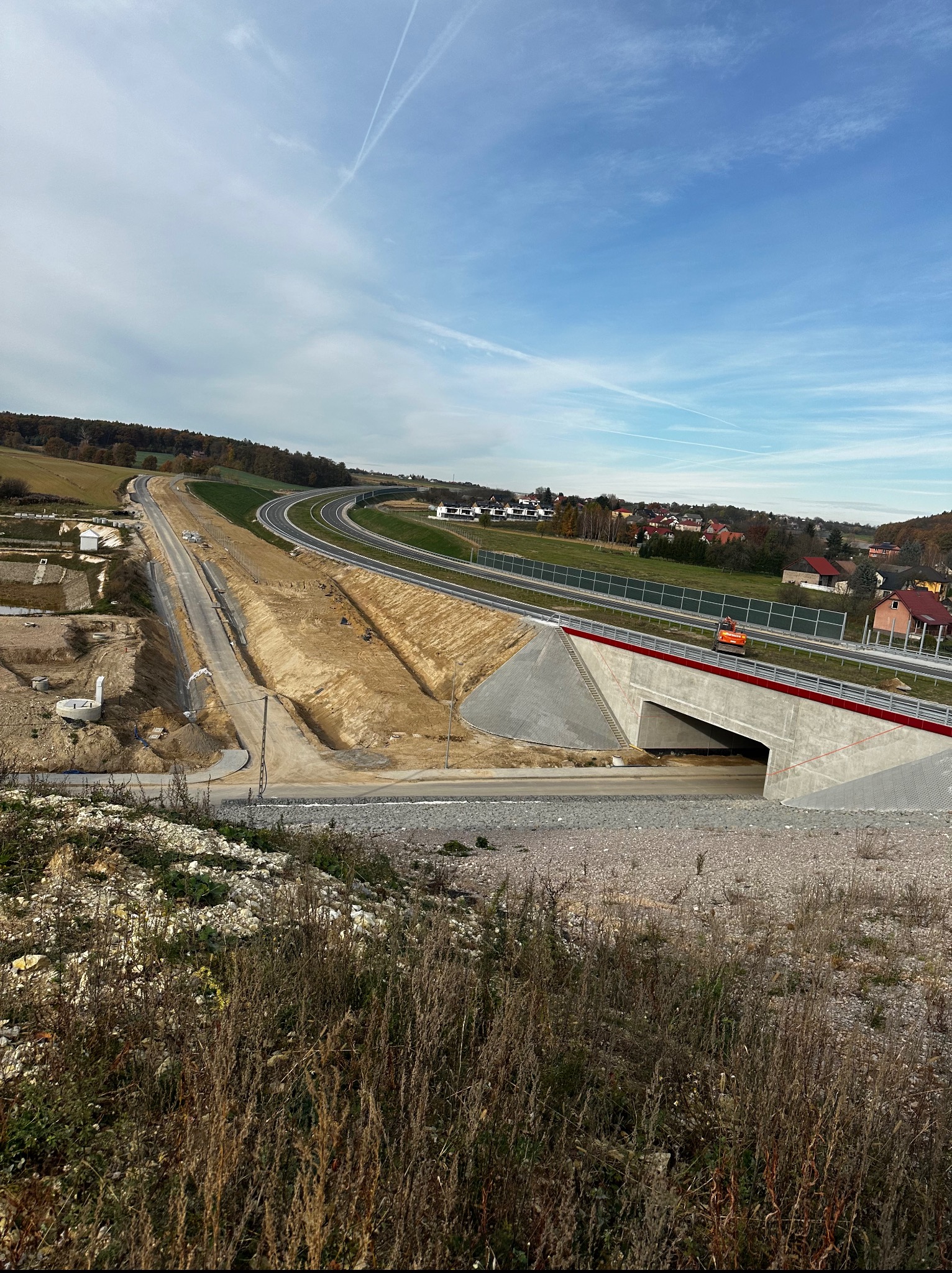 Widok z góry na budowaną drogę ekspresową z tunelem, otoczoną zielenią i zabudowaniami. W oddali widoczne niebo z smugami kondensacyjnymi.
