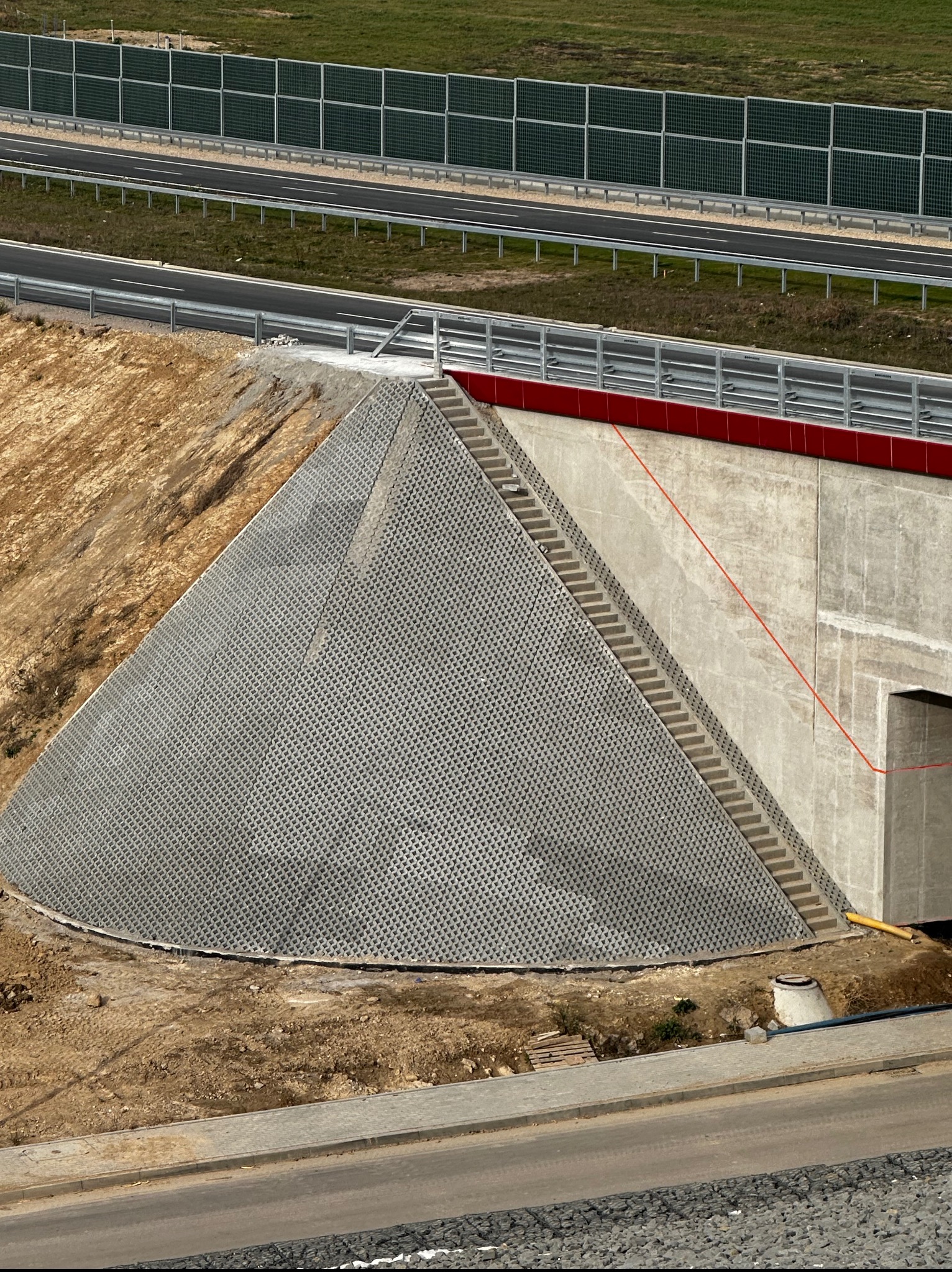 Nowoczesna konstrukcja betonowa z wzorzystym wykończeniem, schodami i czerwoną belką, otoczona ziemią i drogą, z ekranami akustycznymi w tle.
