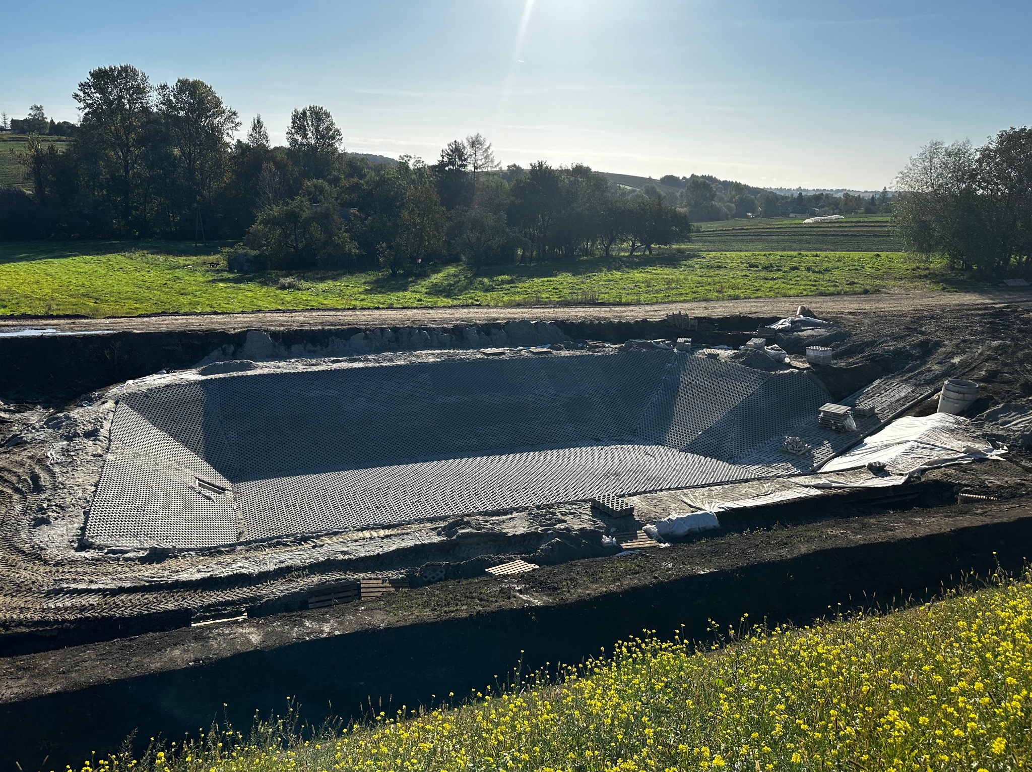 Wyłożony siatką drenażową wykop pod zbiornik wodny na tle zielonego krajobrazu z żółtymi kwiatami. W tle widoczne drzewa i pola uprawne.